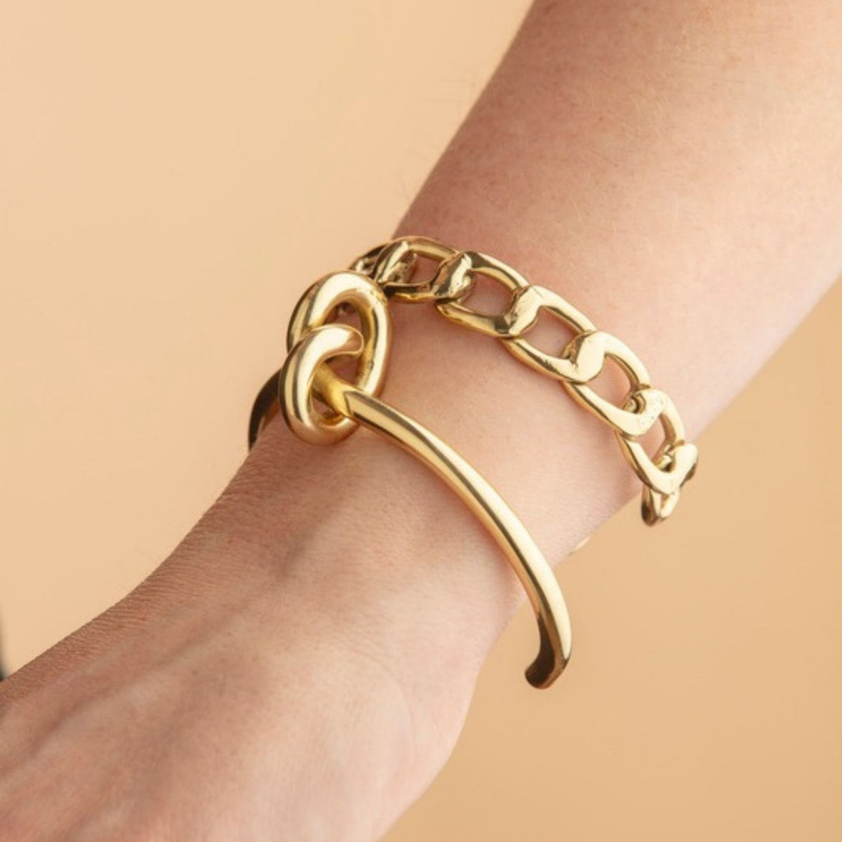 Wrist wearing a gold knot cuff stacked with a chunky gold flat chain link bracelet against a tan backdrop