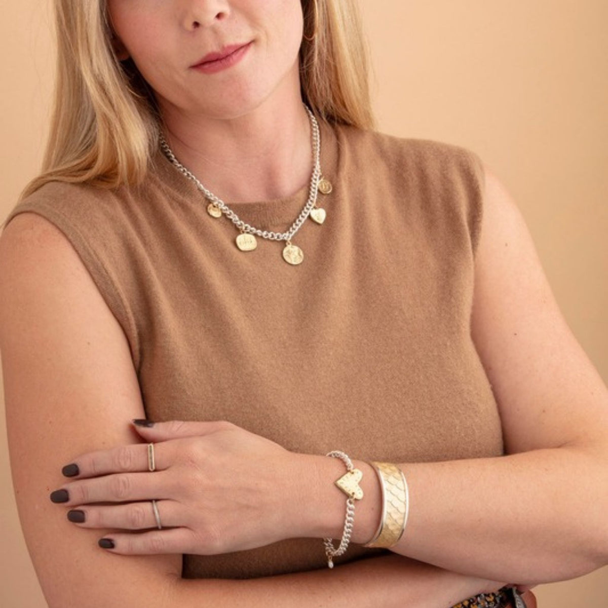 Woman in a camel top with arms crossed wearing a layered charm necklace and a chain bracelet with a gold heart