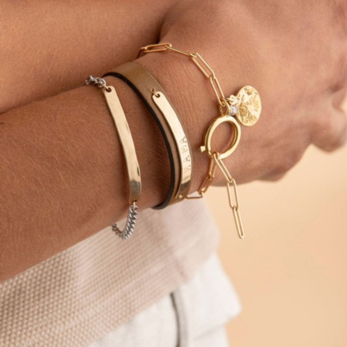 Forearm wearing a gold engraved bar bracelet, a leather cuff and a paperclip chain bracelet with a coin charm