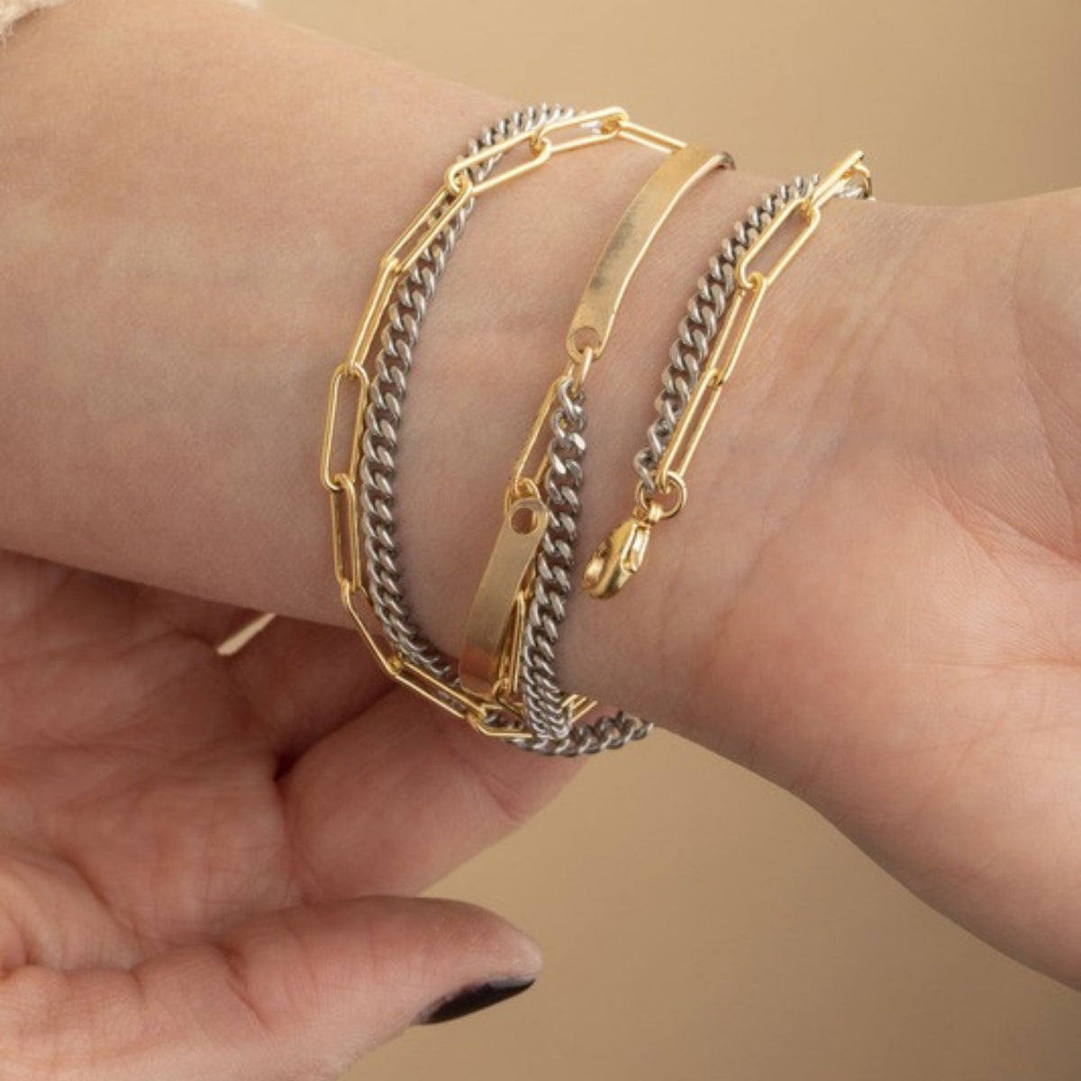 Wrist stack of gold paperclip chain, silver curb chain and a gold ID bar bracelet with a lobster clasp