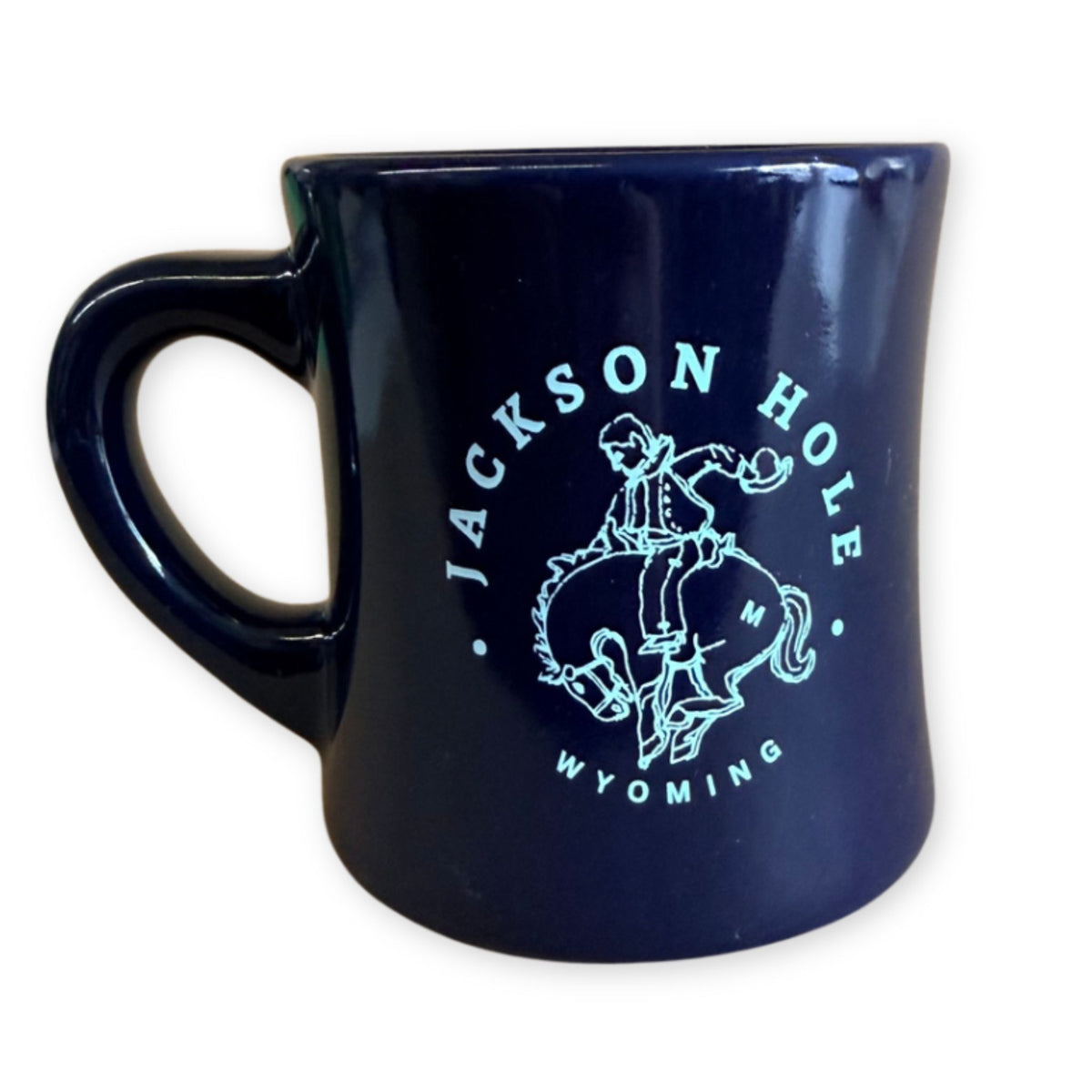 A glossy dark navy blue ceramic mug featuring a light blue graphic of a cowboy on a bucking bronco with the text 'JACKSON HOLE WYOMING' printed around it.