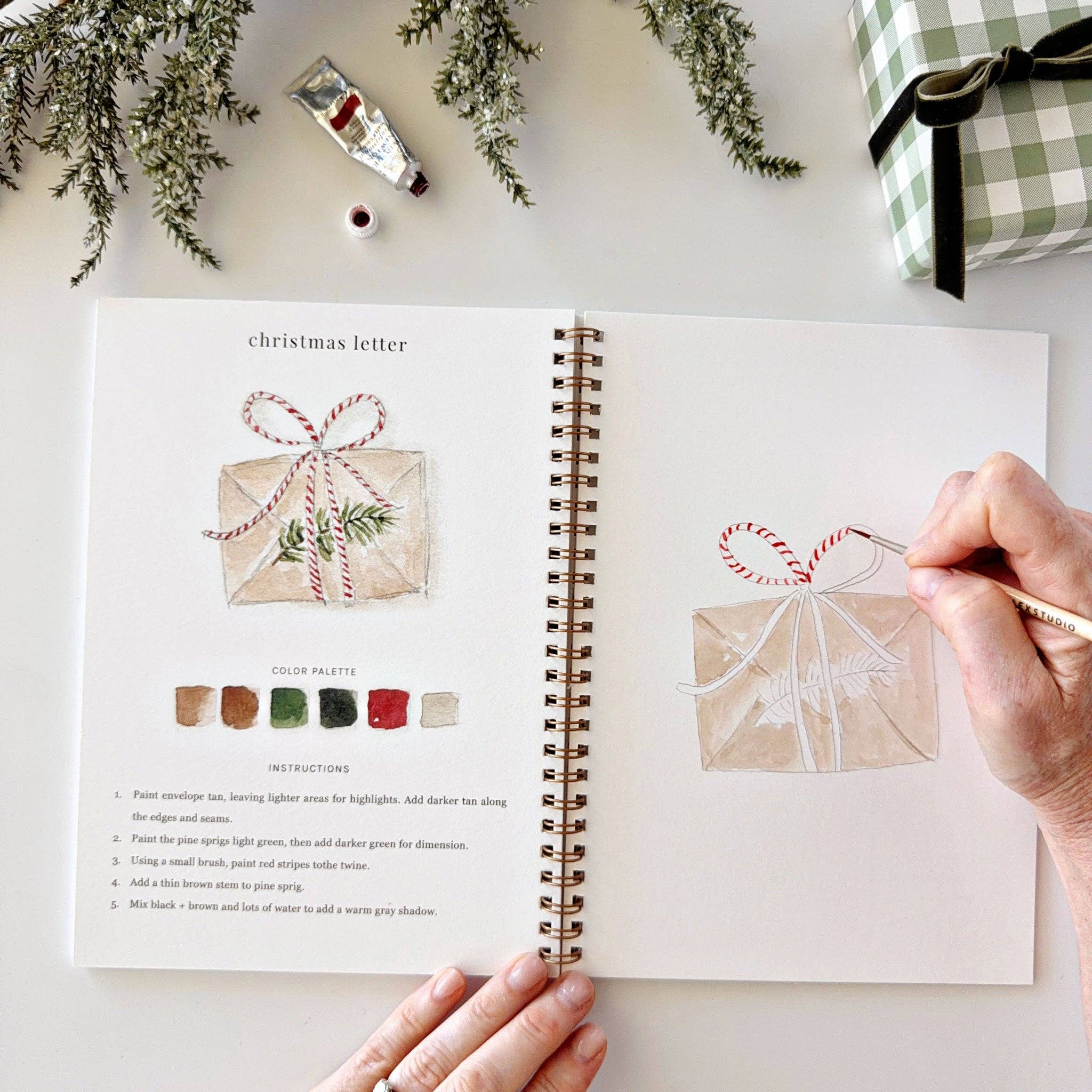 An open spiral-bound art journal shows a Christmas letter painting instruction page with illustrated wrapped gifts and color swatches. A person's hands are visible working on painting a matching illustration in the journal, with holiday greenery and a wrapped gift in the background.