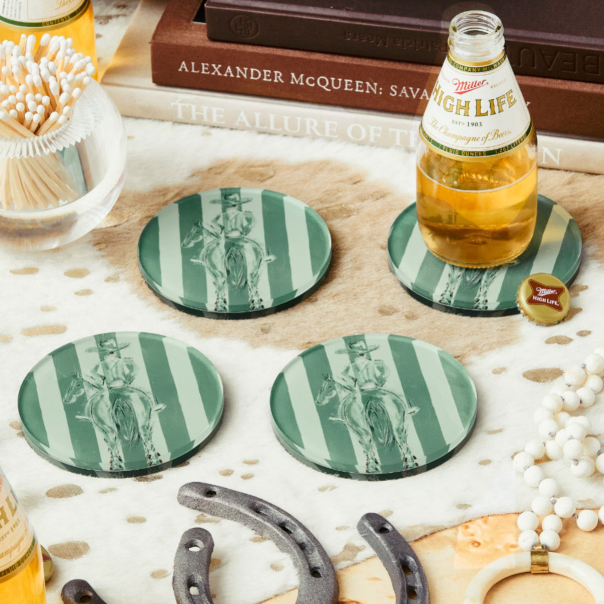 Four round glass coasters with a green and white striped design featuring a vintage cowboy illustration, arranged on a rustic tabletop with books, a bottle of Miller High Life, horseshoes, and other decorative props.