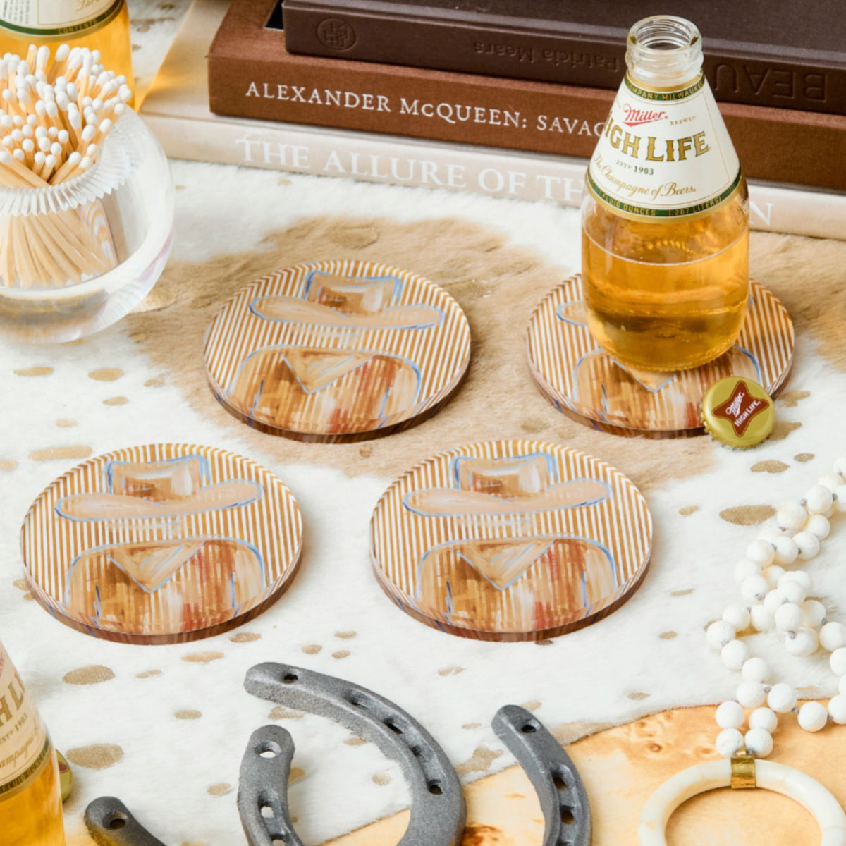 Four rectangular wooden coasters with a vintage beer bottle illustration arranged on a styled tabletop with books, a glass bottle of beer, a bottle cap, horseshoes, and decorative beads.