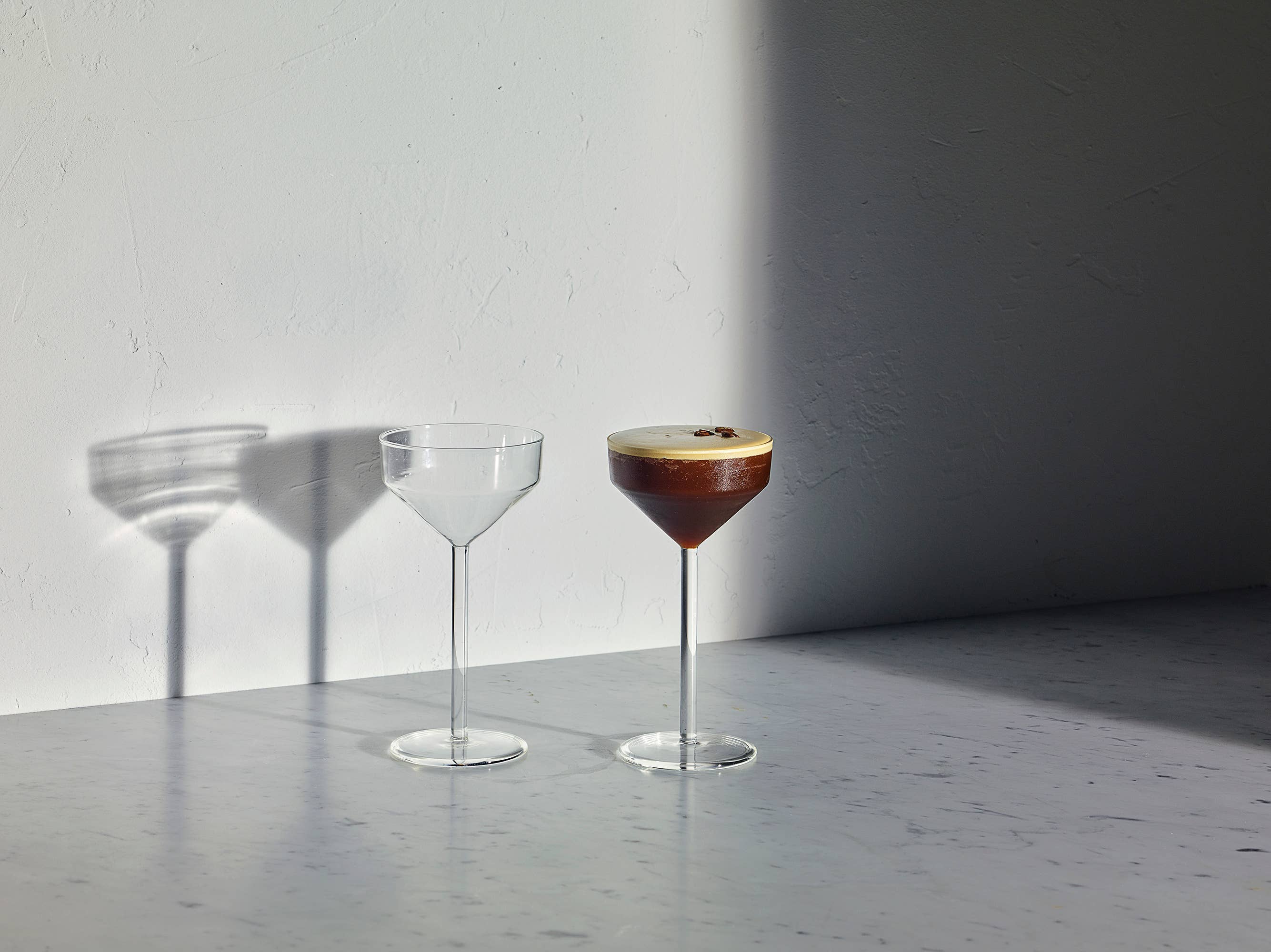 Two clear stemmed cocktail glasses on a gray surface against a white wall, with one glass empty and the other filled with a dark brown drink topped with cream, casting long shadows.