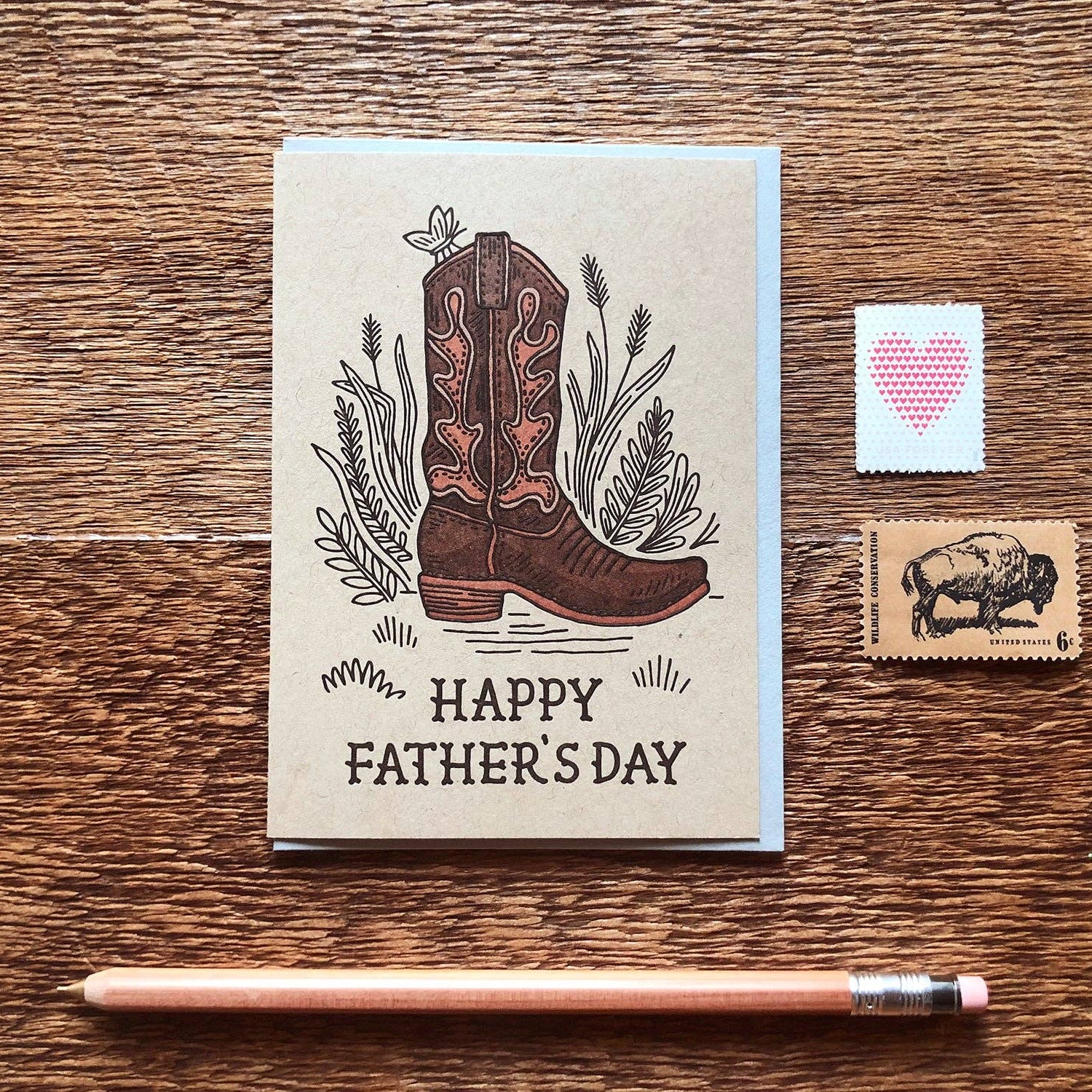Card with cowboy boot illustration and 'Happy Father's Day' text on a wooden surface with stamps and a pencil.