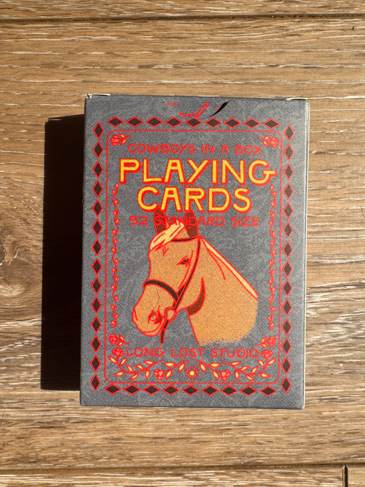 A vintage-style cowboy playing cards deck box. The box is gray with a red border and features an illustration of a cowboy horse head.