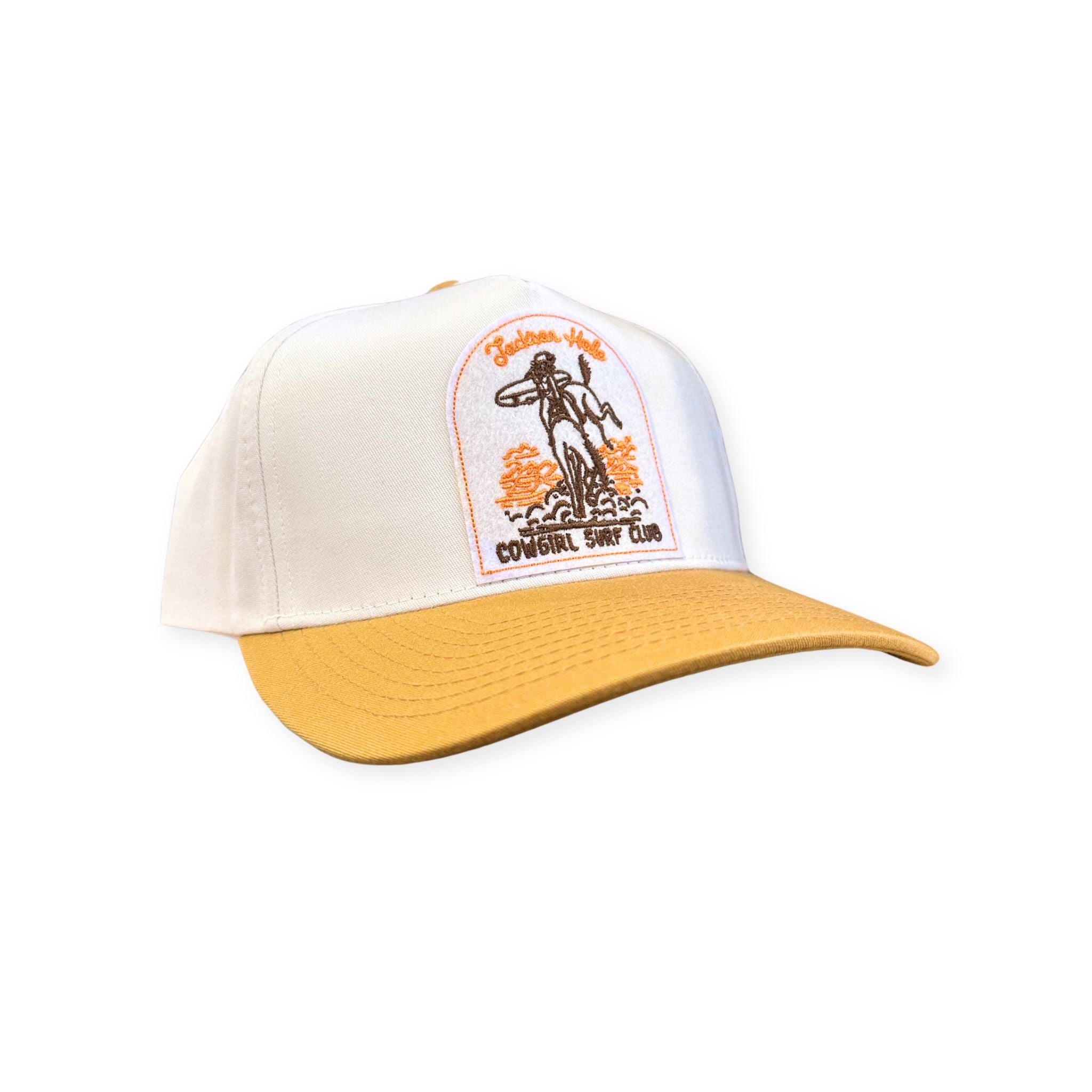 A two tone trucker hat. Colors are a yellowy tan, and white. Featuring the cowgirl surf club patch in the middle. Colors of the patch are white, orange, and brown.