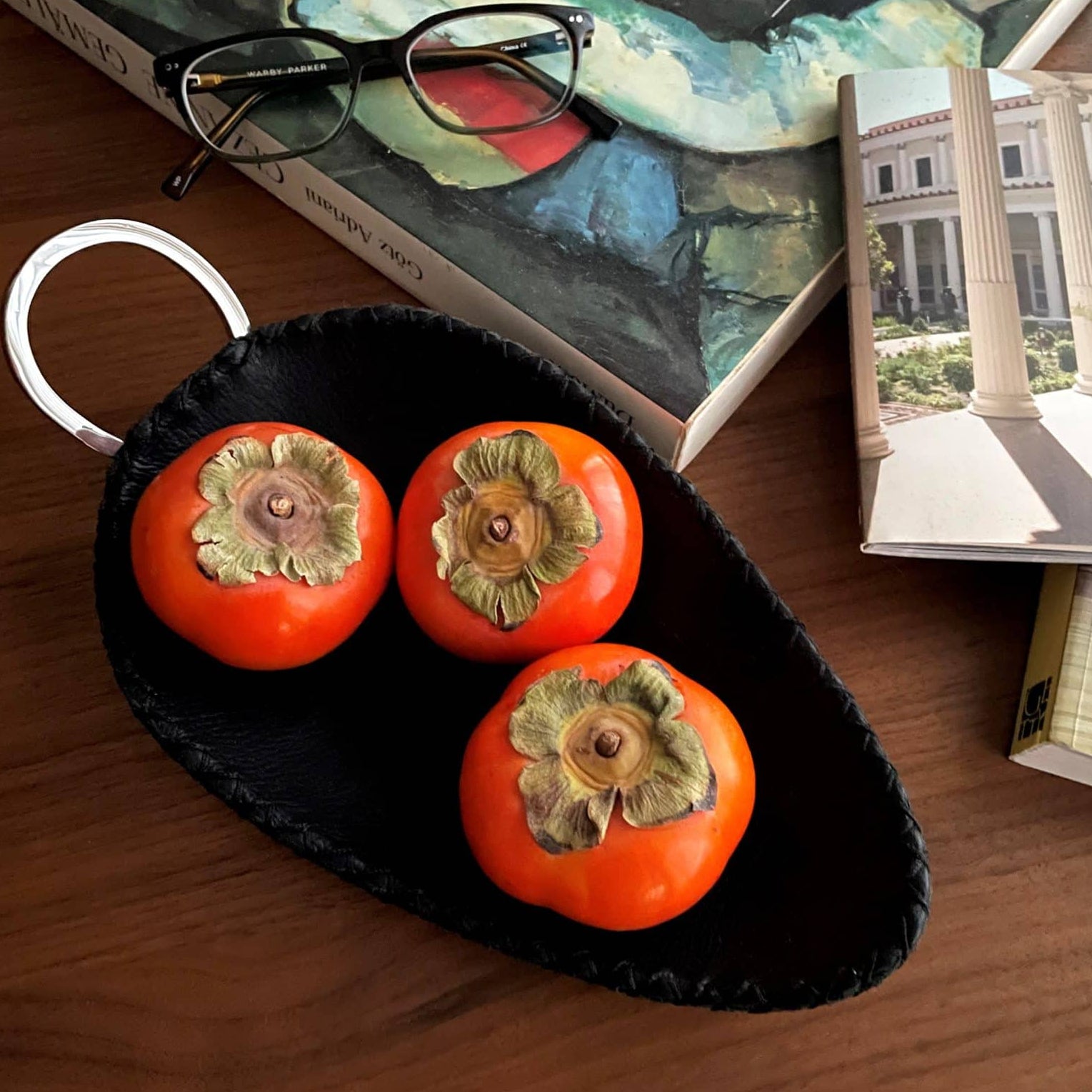 Three persimmons in a black dish on a wooden surface with books in the background