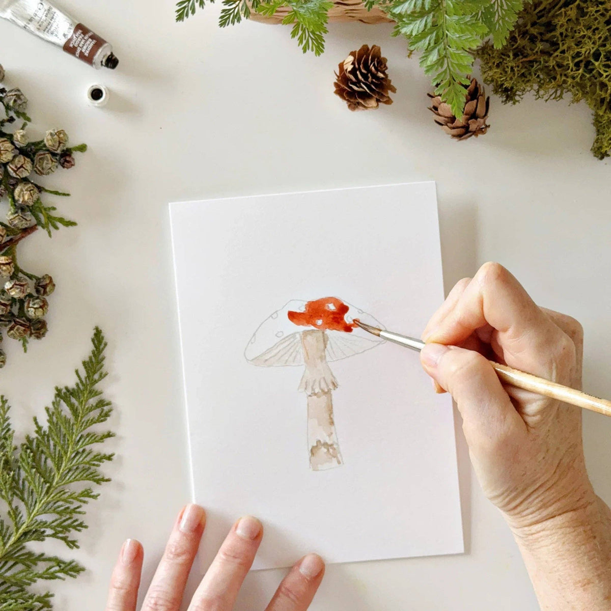 Person painting a small figurine on a white card with greenery and pinecones around