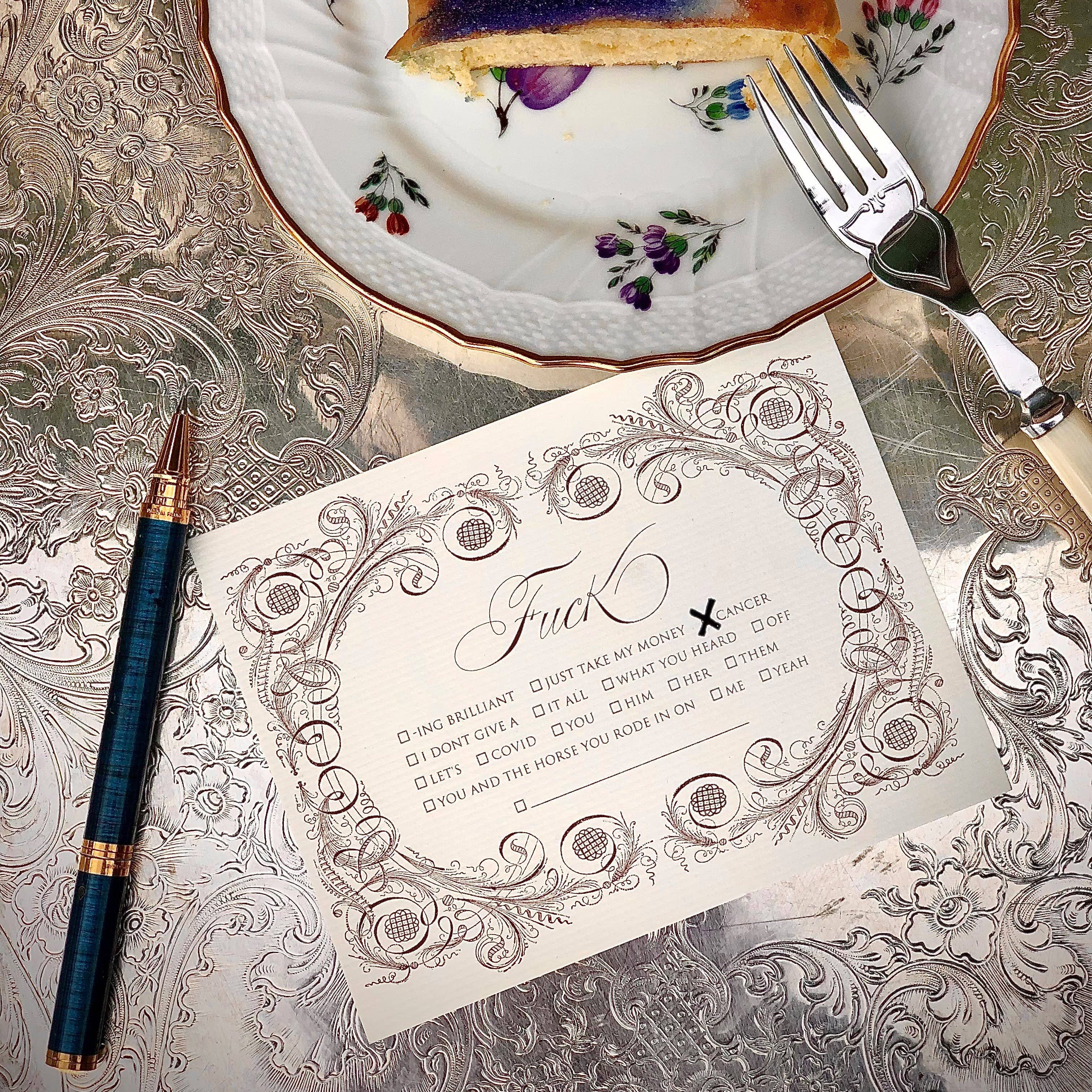 Slice of cake on a decorative plate with a fork, pen, and ornate card on a patterned surface.