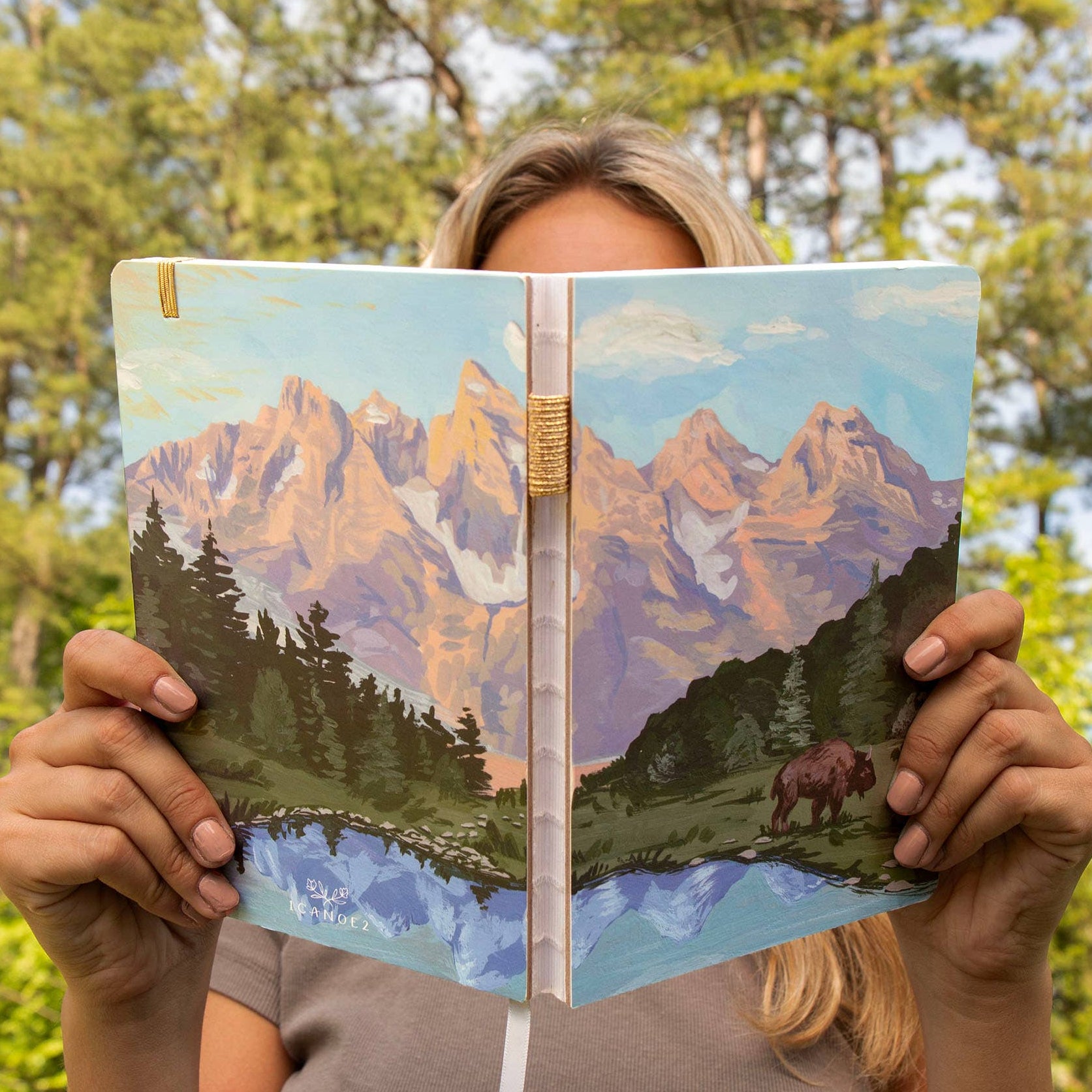 Person holding a notebook with a mountain and bison illustration in a forest setting