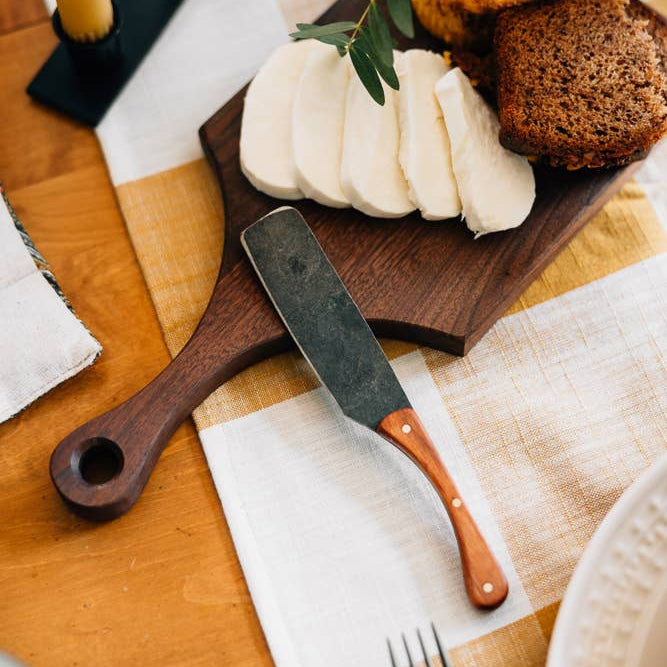 Wooden cutting board with bread, cheese, and a knife on a table setting.