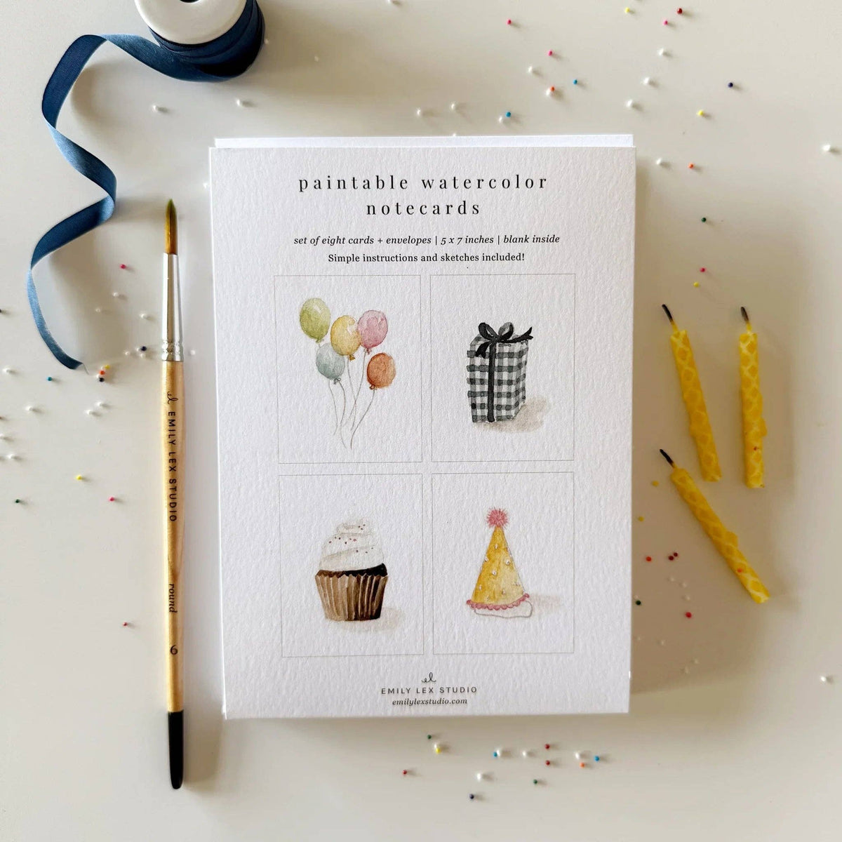 A flat-lay of a card set package labeled 'paintable watercolor notecards' on a light beige surface, surrounded by a paintbrush, blue ribbon, and yellow candles. The package shows four watercolor illustrations: balloons, a gift box, a cupcake, and a party hat.