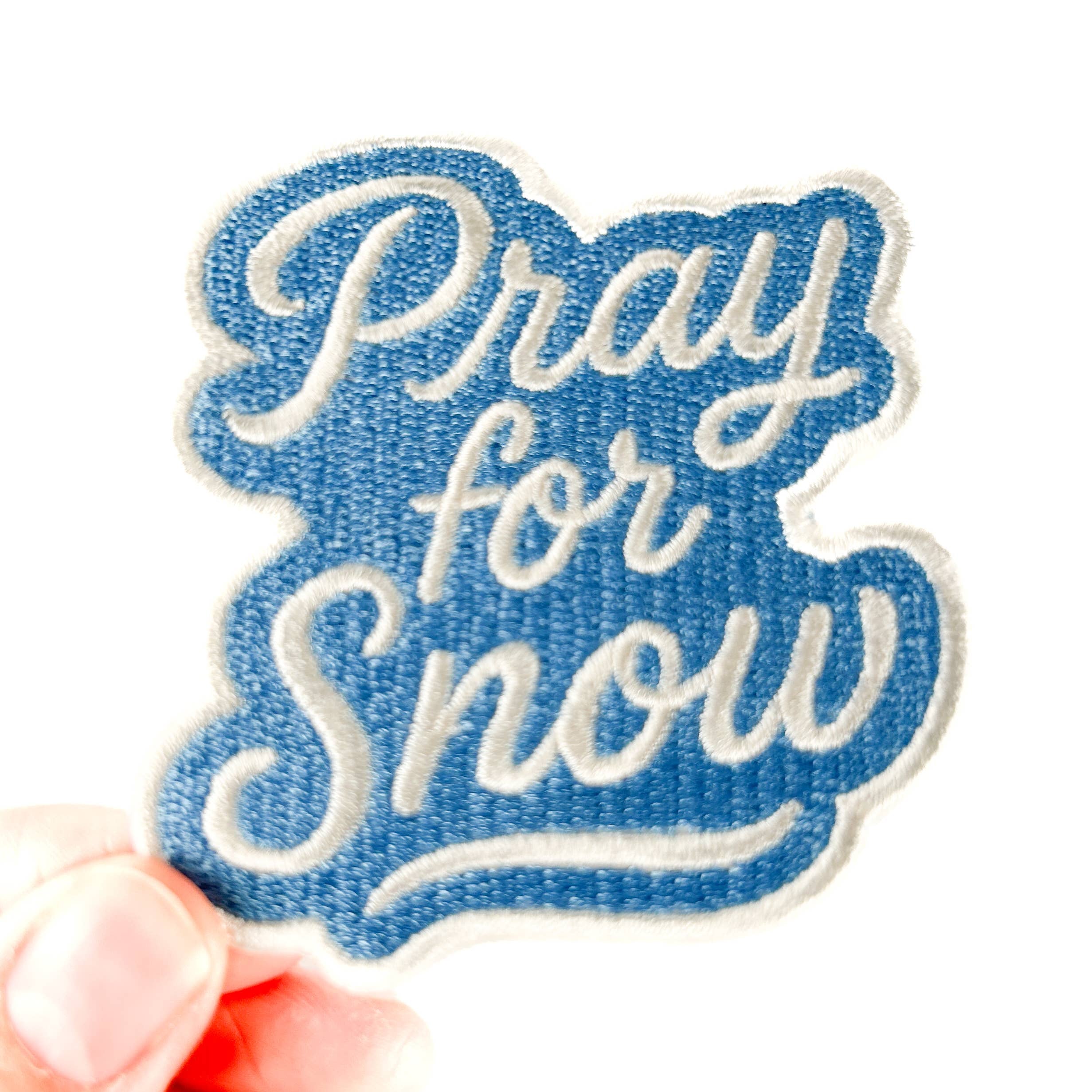 Denim patch with 'Pray for Snow' text held by a hand on a white background
