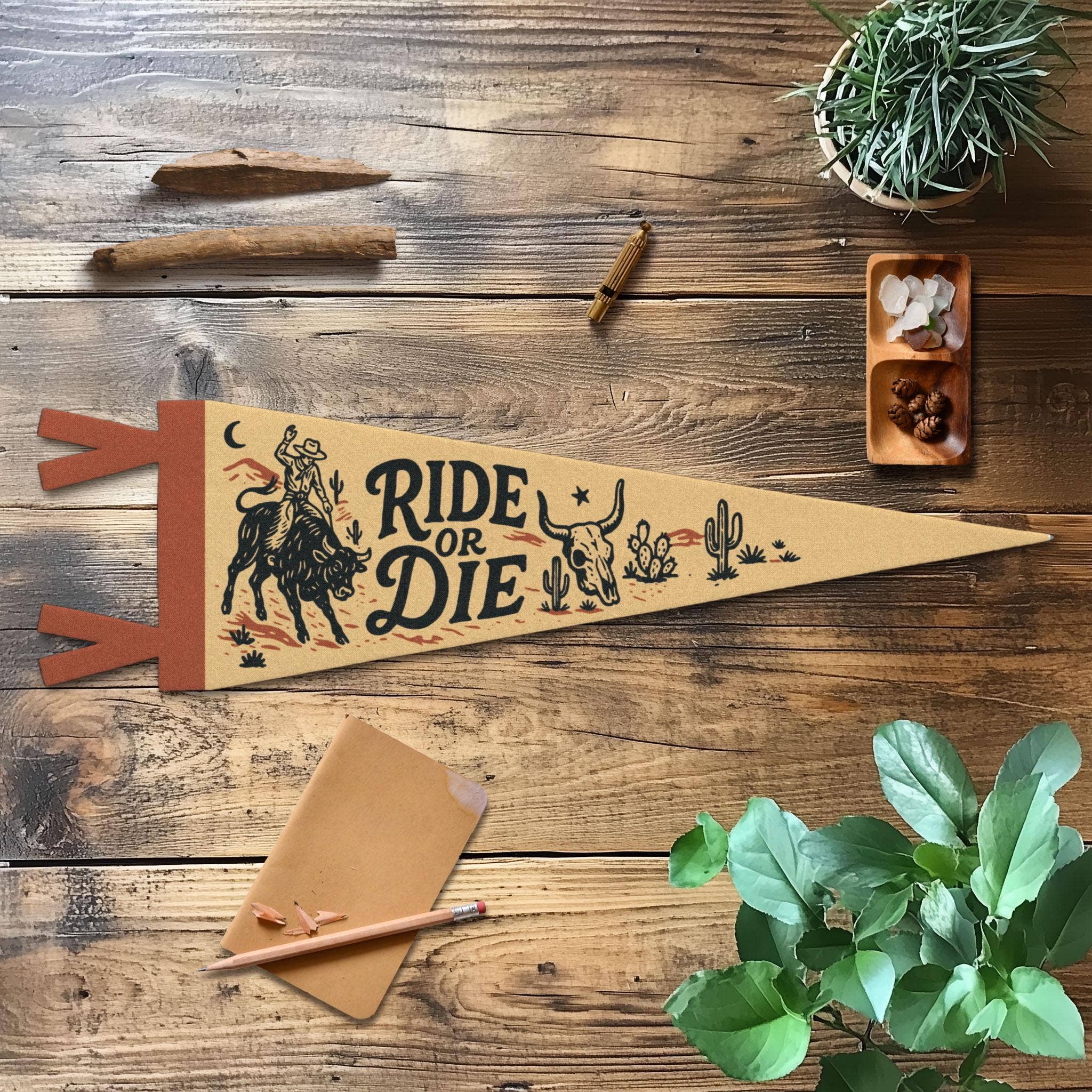 Decorative pennant with 'Ride or Die' text on a wooden surface with plants and stationery.