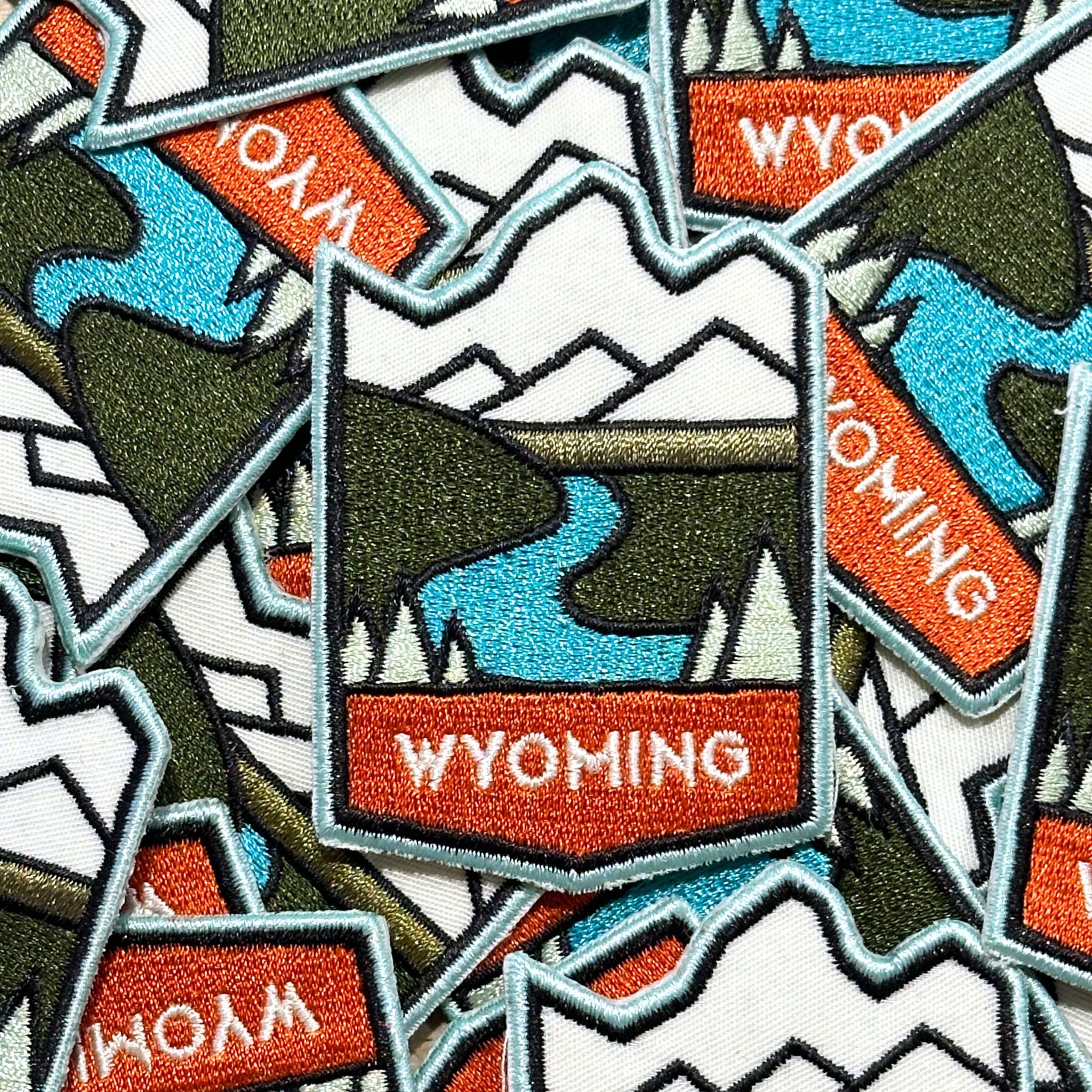 Embroidered patch with marrowed edge. Showing the winding snake river, mountains, and trees. With a curved top and diamond shape bottom. Wyoming is written in white all capital lettering against an orange back.