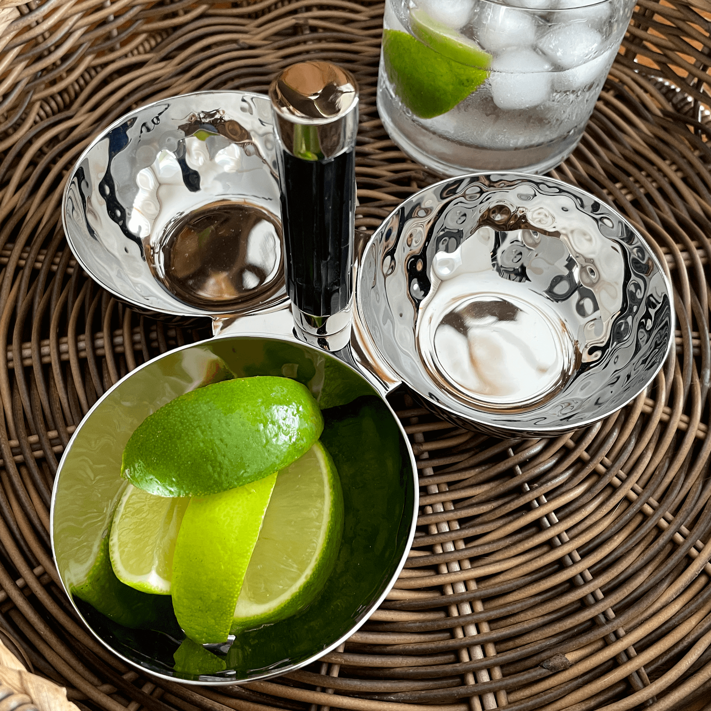 Bar tools and ingredients on a woven mat with lime wedges and a glass of ice.