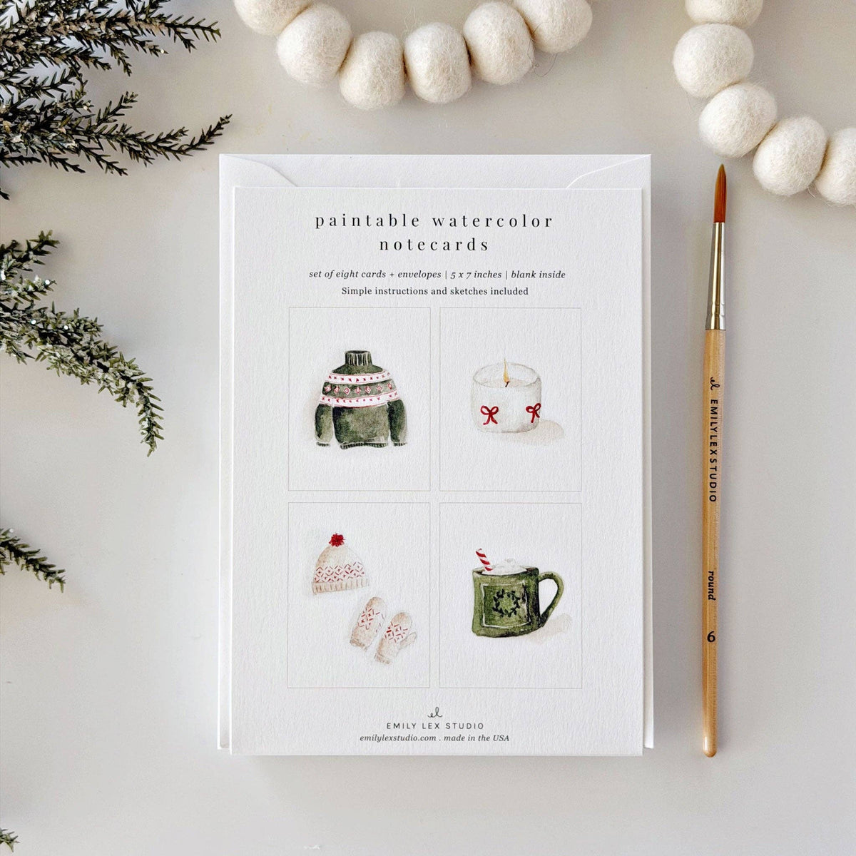 A flat-lay of a notepad card titled 'paintable watercolor notecards' showing four small watercolor illustrations, styled with white felt balls, pine branches, and a paintbrush on a light background.