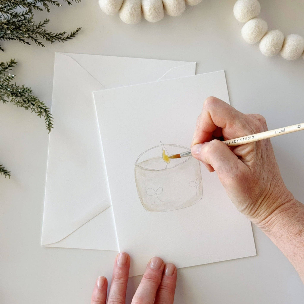 Painting a candle jar on a white card layered over a white envelope, with felt garlands and greenery in the background.
