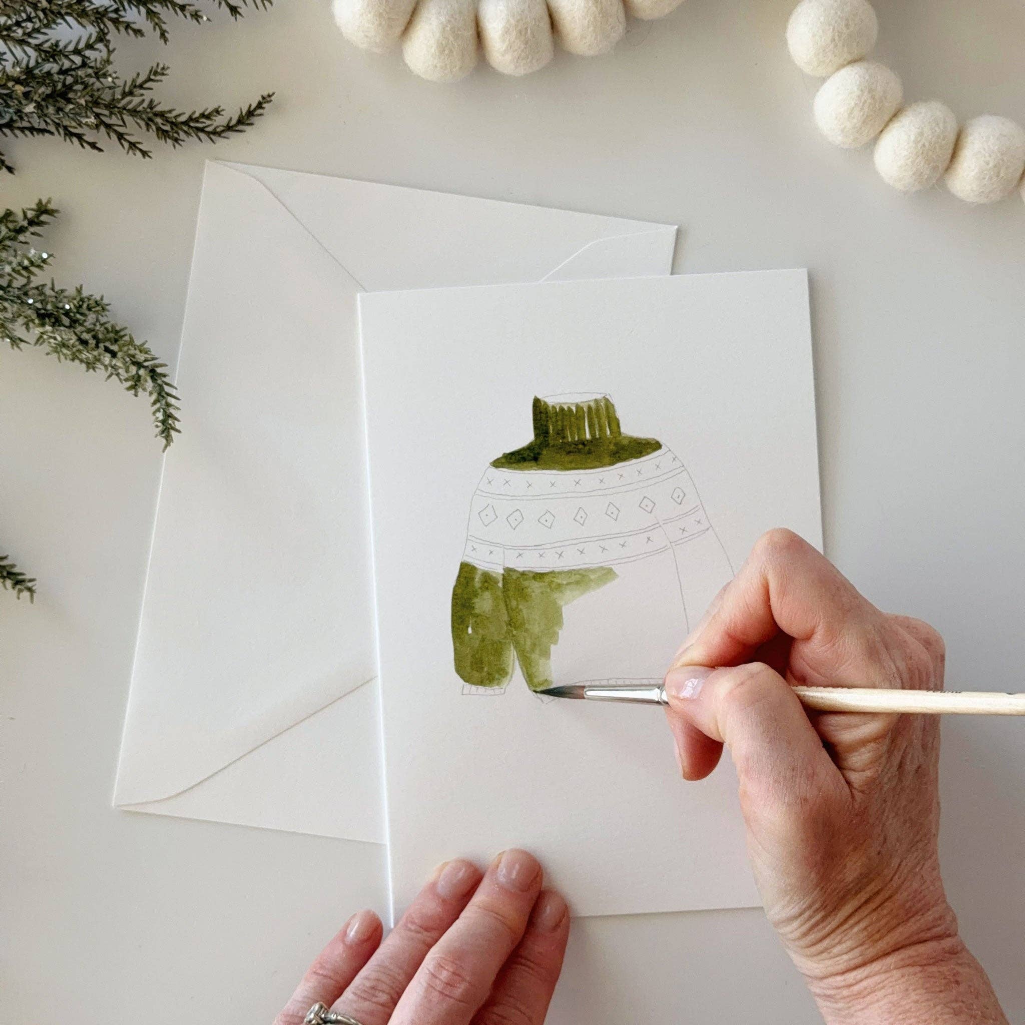 A close-up of a hand holding a paintbrush and painting an olive green illustration of a Santa Claus hat on a white card, with an envelope behind it on a light tabletop.