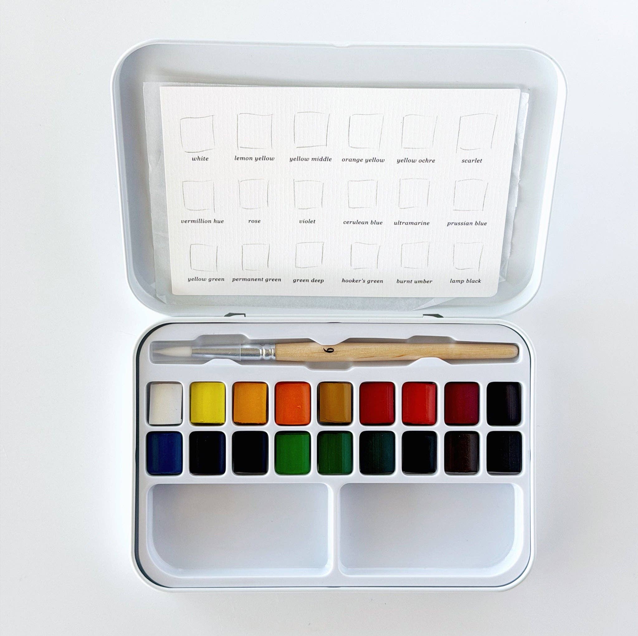 An open metal watercolor paint set with a white tray holding multiple color pans, a wooden-handled brush, and a sheet of color cards inside the lid.