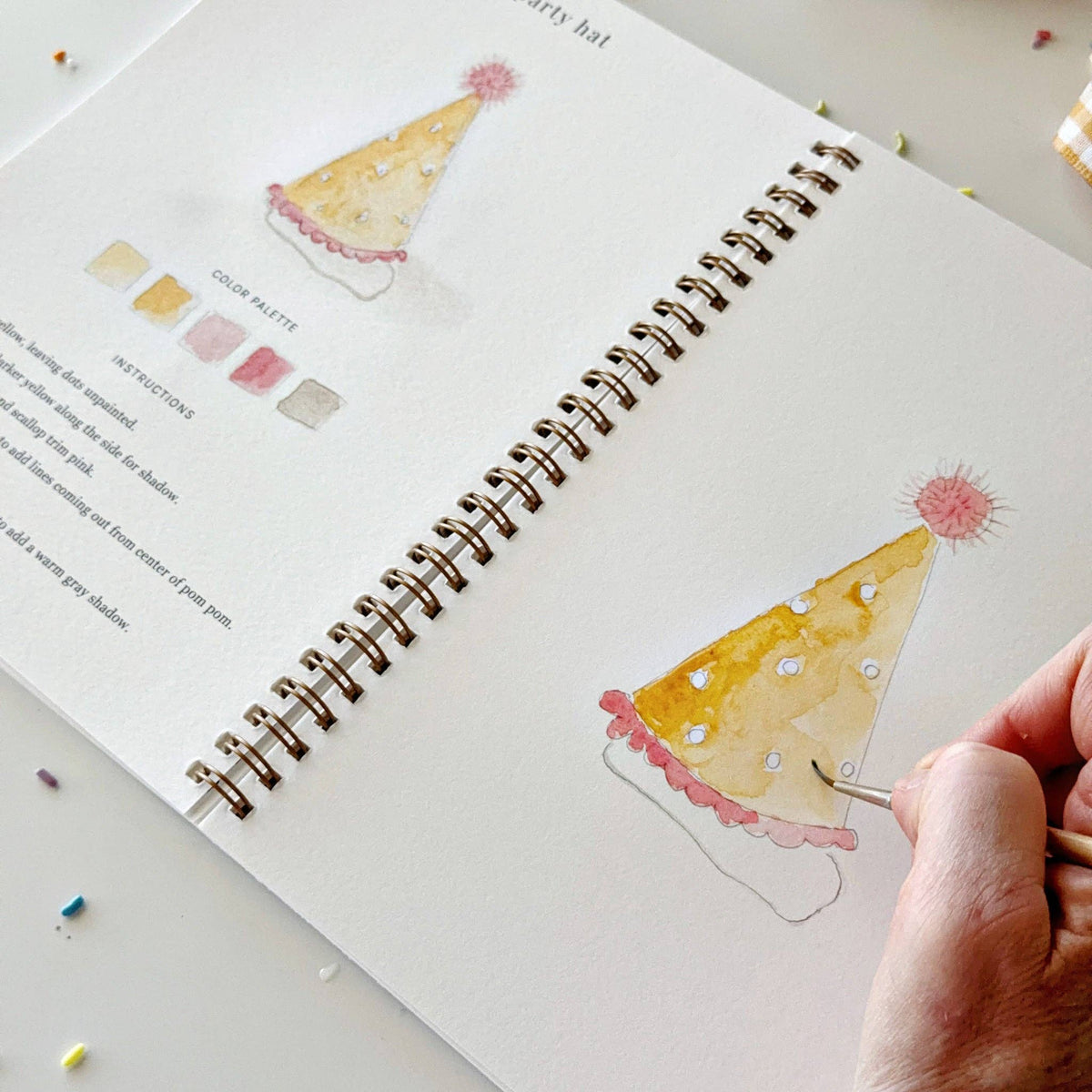 Person drawing a party hat illustration in a sketchbook with watercolor techniques.