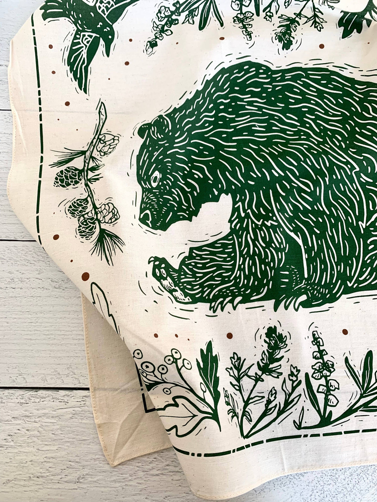 A cream-colored Bandana featuring a dark green hand-drawn style illustration of a bear, pinecones, and wildflowers.