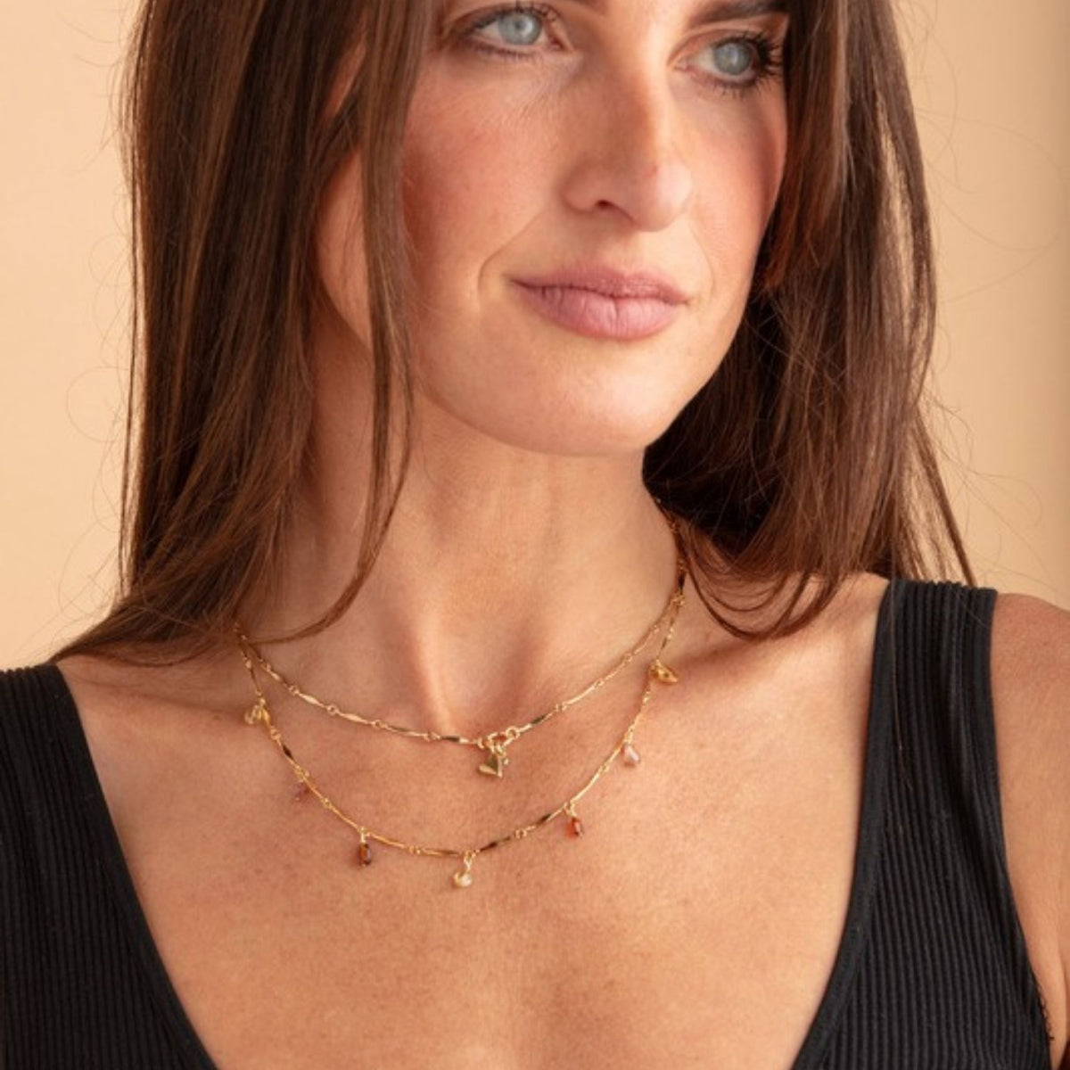 Woman in a black ribbed tank top wearing two layered gold bar-chain necklaces with small heart and drop charms