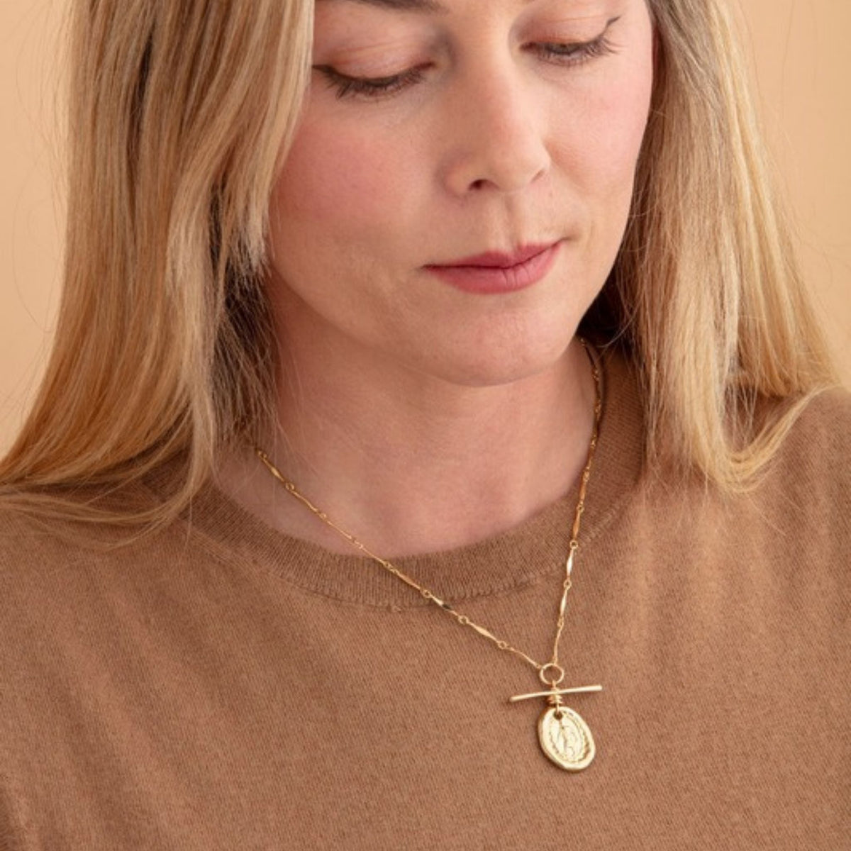 Woman in a camel knit top looking down, wearing a gold bar-chain necklace with a T-bar and oval medal