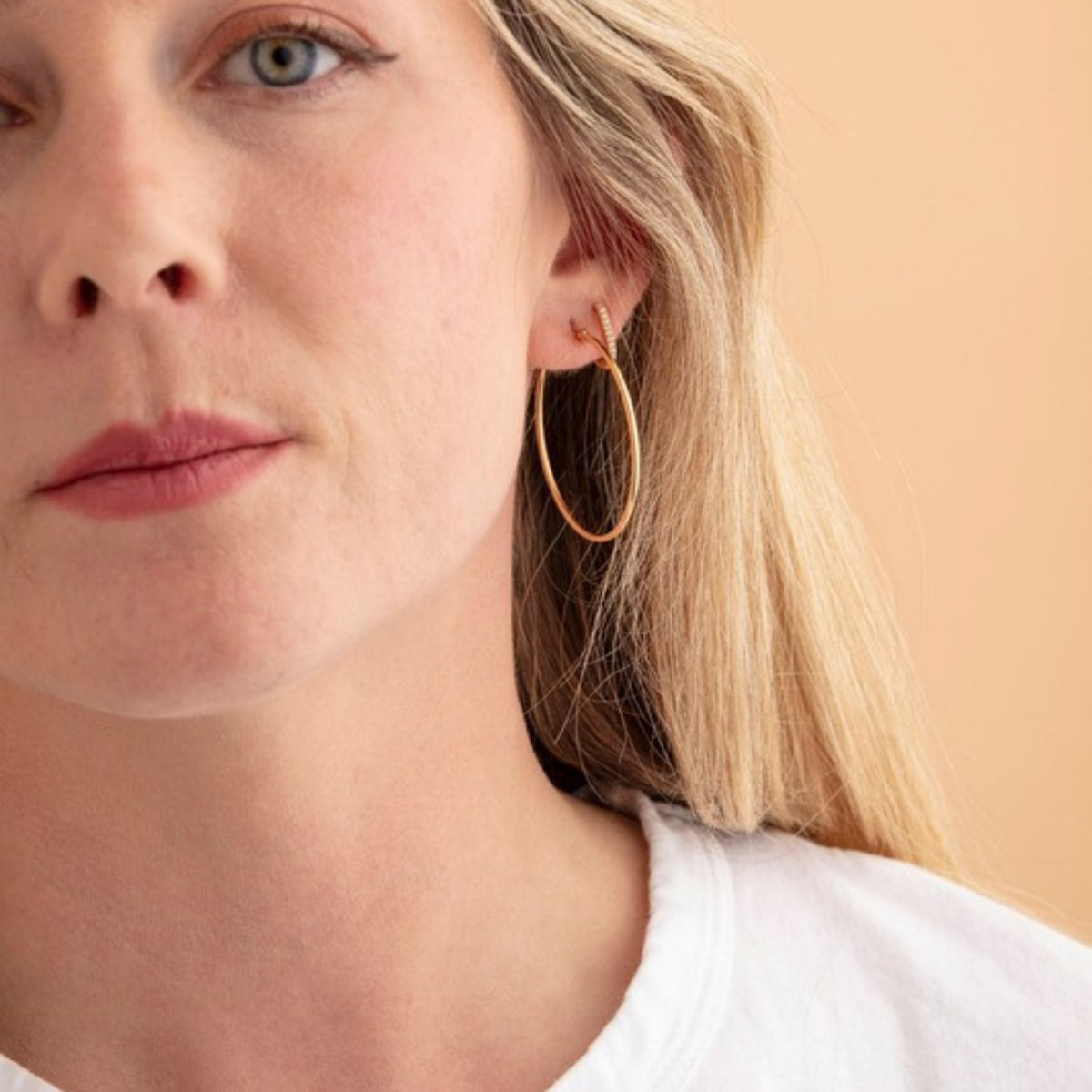 Close crop of a woman's face and ear wearing a thin gold hoop earring, white tee at the collar