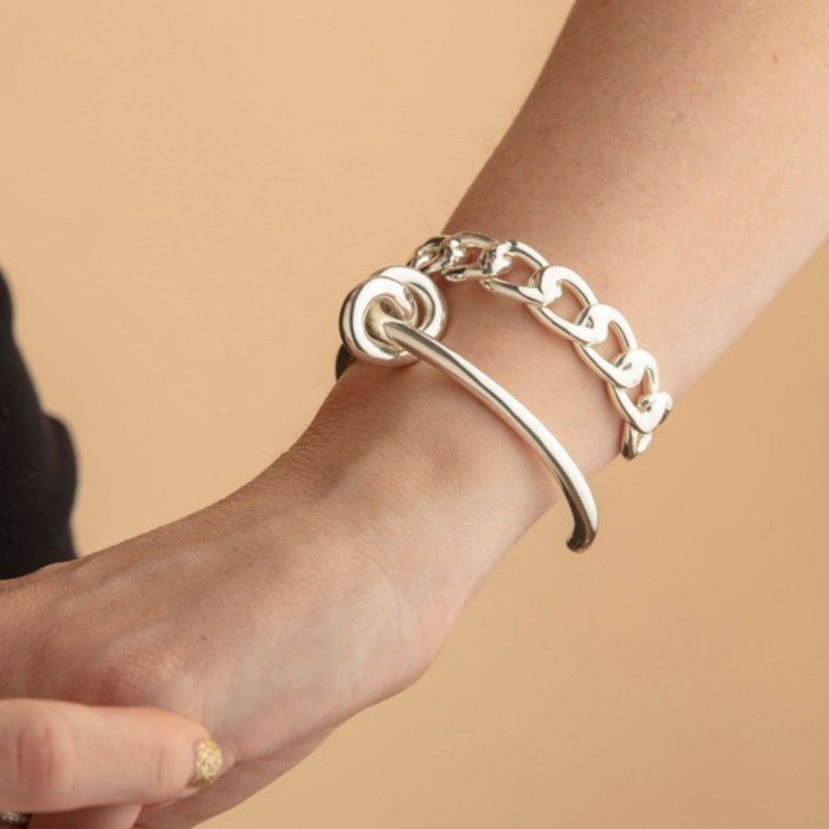 Wrist wearing a silver knot cuff stacked with a chunky silver flat chain link bracelet against a tan backdrop
