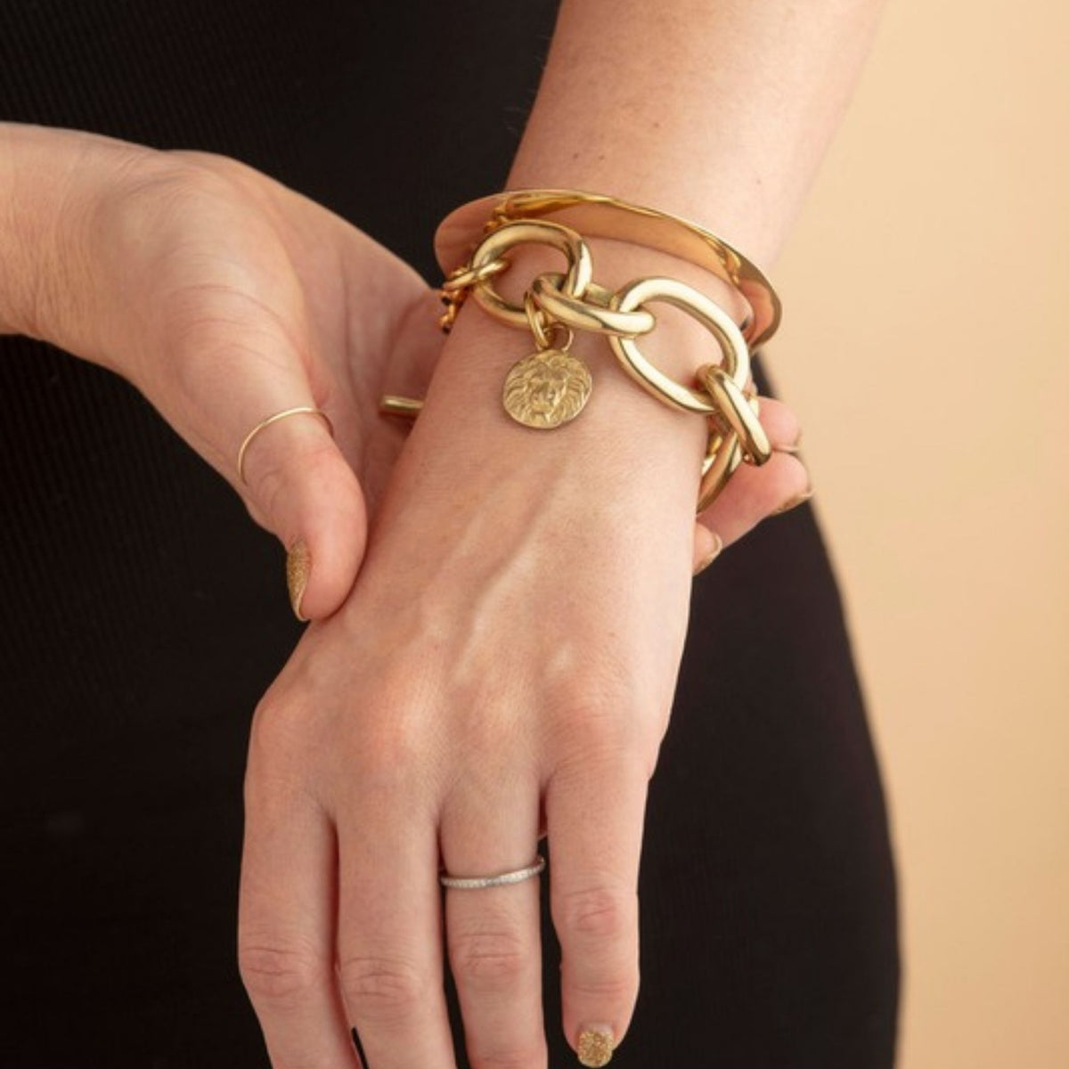 Hands crossed in front of a black top wearing a gold tapered cuff stacked with a chunky oval chain bracelet and coin charm