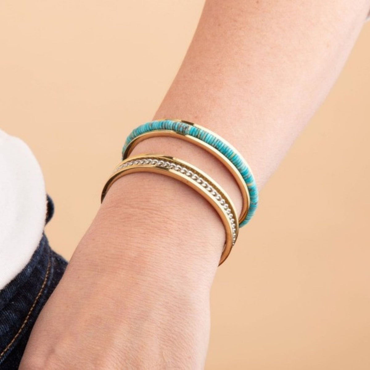 Wrist wearing a gold cuff with turquoise disc beads stacked with a gold cuff with a silver chain inlay