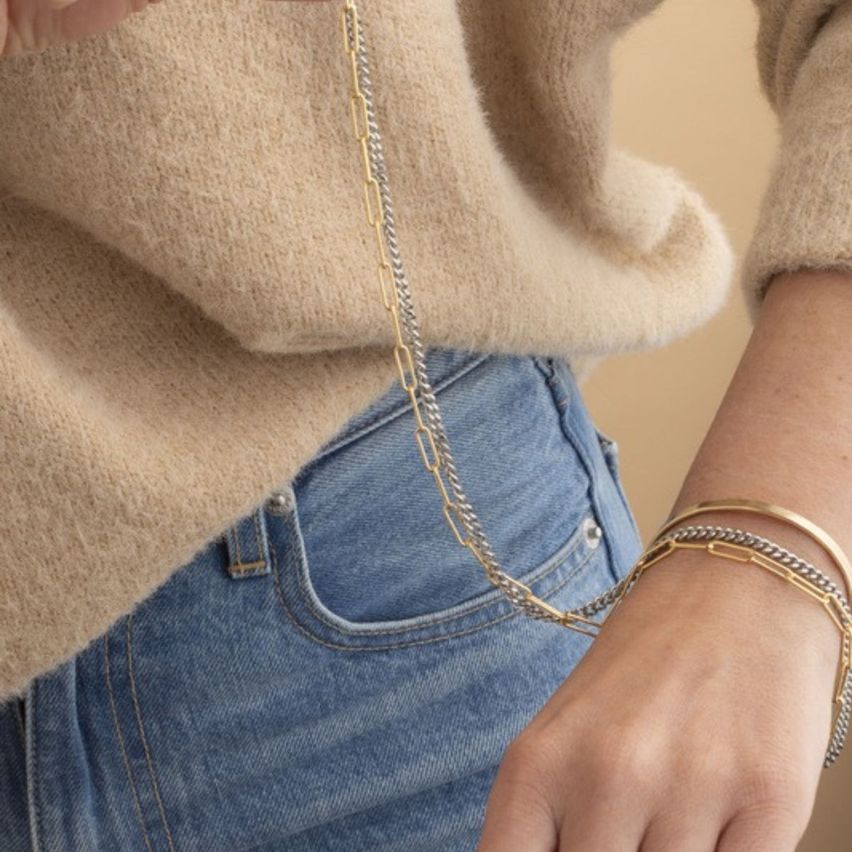 Woman in a fuzzy tan sweater and jeans holding a long mixed gold and silver chain over stacked bracelets