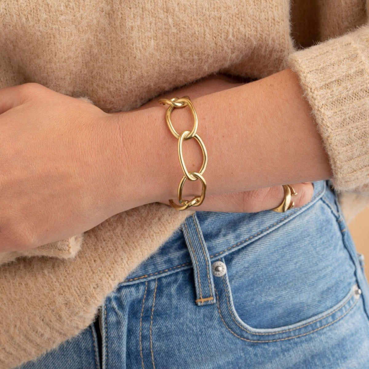 Wrist in a tan sweater and jeans wearing a gold oval open-link chain bracelet with a looped clasp detail