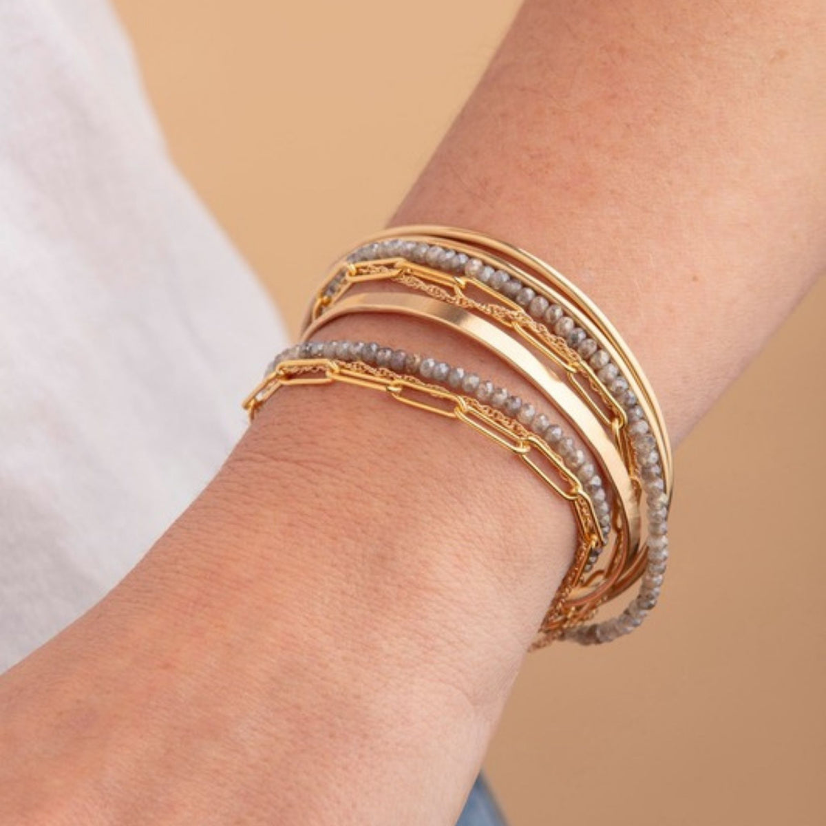 Wrist wearing a stack of gold bangles, paperclip chain and gray bead bracelets beside a white tee sleeve