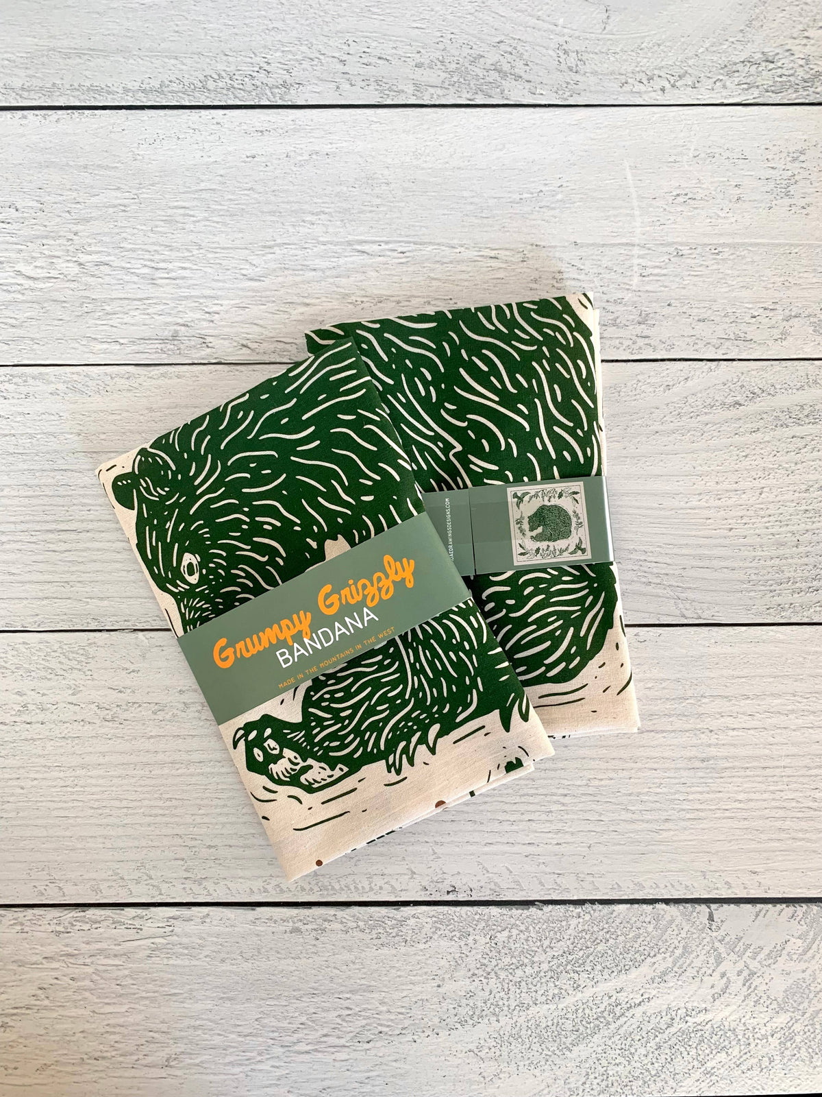 Two folded cotton bandanas with a dark green, pine pattern, wrapped in a green paper band labeled 'Grumpy Grizzly Bandana' and a paper sleeve.