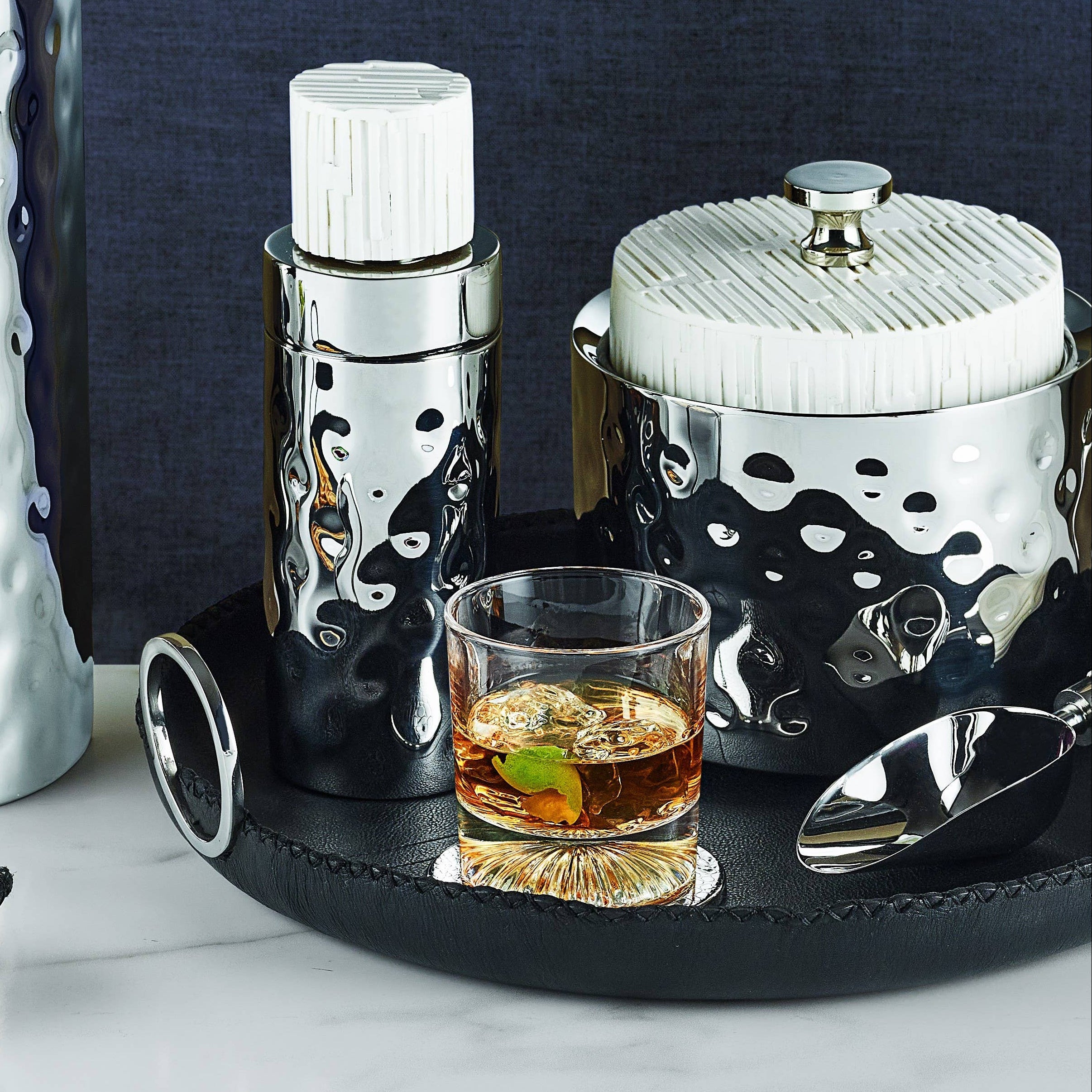 Set of cocktail-making tools including a shaker, strainer, and glass with a drink on a tray against a dark background.
