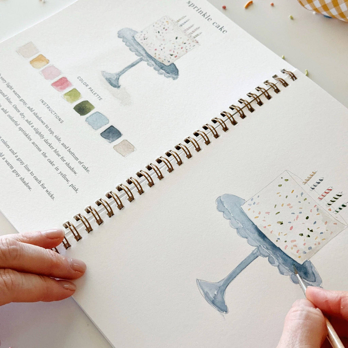 Open notebook with illustrations of a cake stand and color swatches, held by a person.