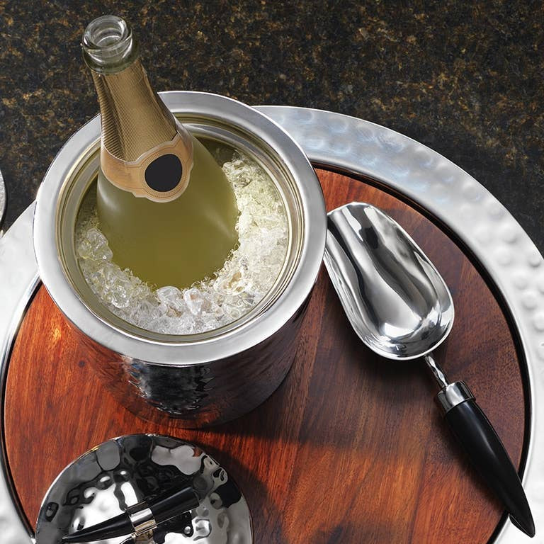 Bottle of champagne in a metal ice bucket with wooden base and serving tools on a dark surface
