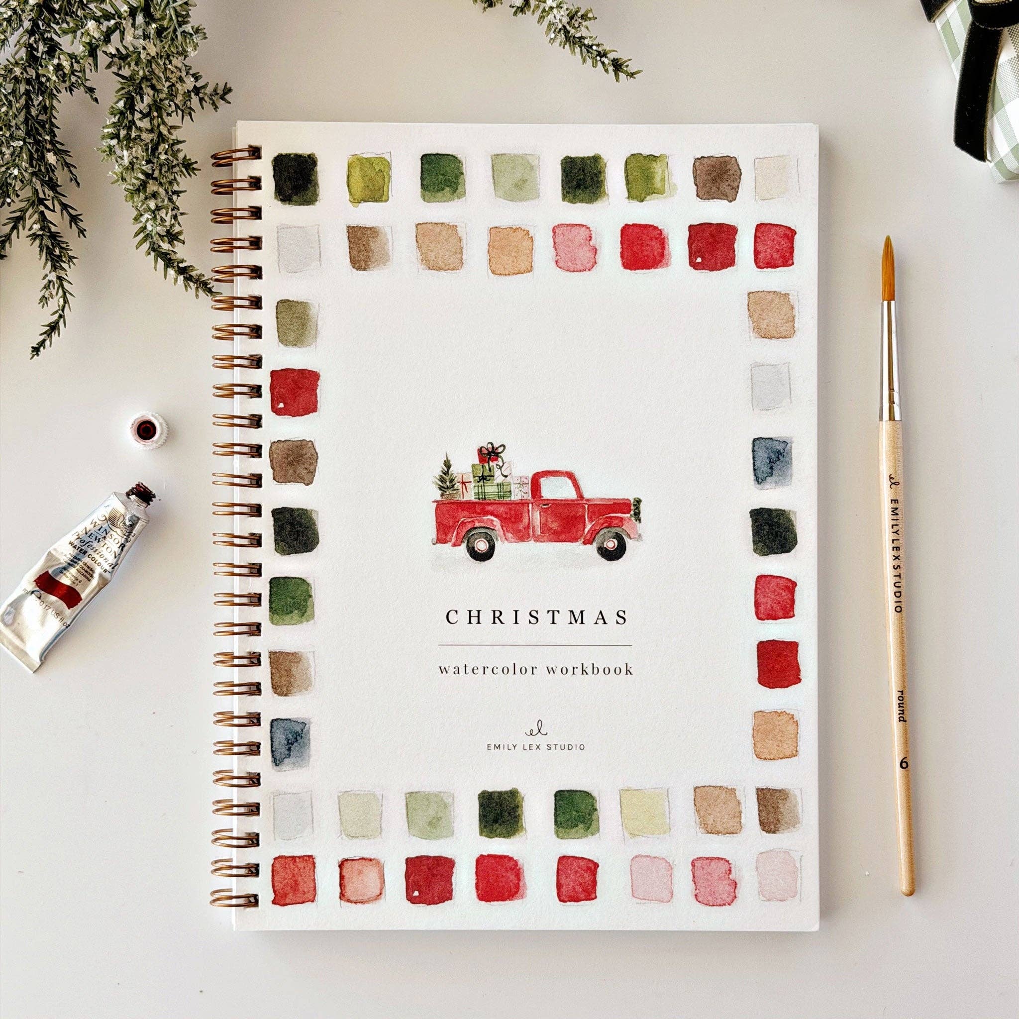A spiral-bound watercolor workbook titled 'CHRISTMAS watercolor workbook' by Emily Lee Studio, styled in a flat lay with a red truck illustration and watercolor swatches around the border. A paint tube, paintbrush, and greenery are arranged around it on a light background.