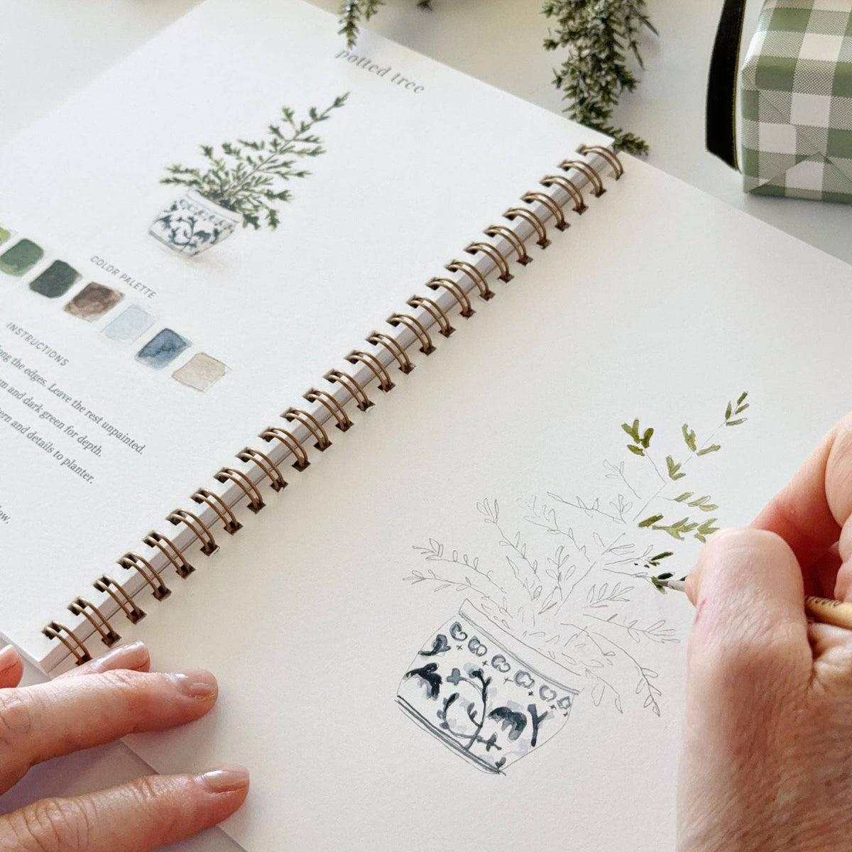 A hand-drawn planner or journal page featuring a potted tree illustration, held open by two hands. The page has a spiral binding and is shown on a light surface with a small green-and-white checkered box in the background.