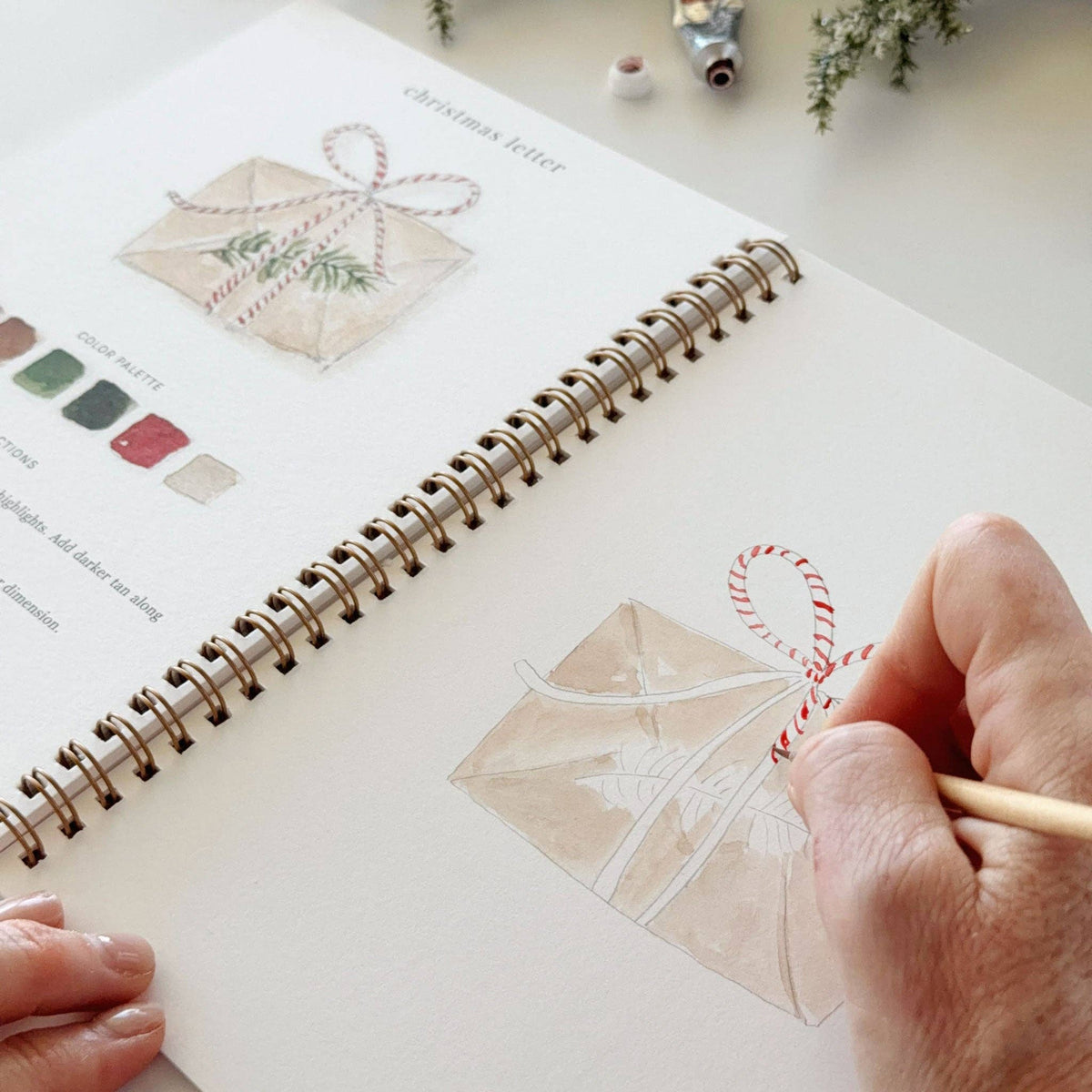 A close-up of a hand drawing a Christmas letter envelope with red and white ribbon on a spiral-bound art journal, with color swatches and another illustration visible on the page.