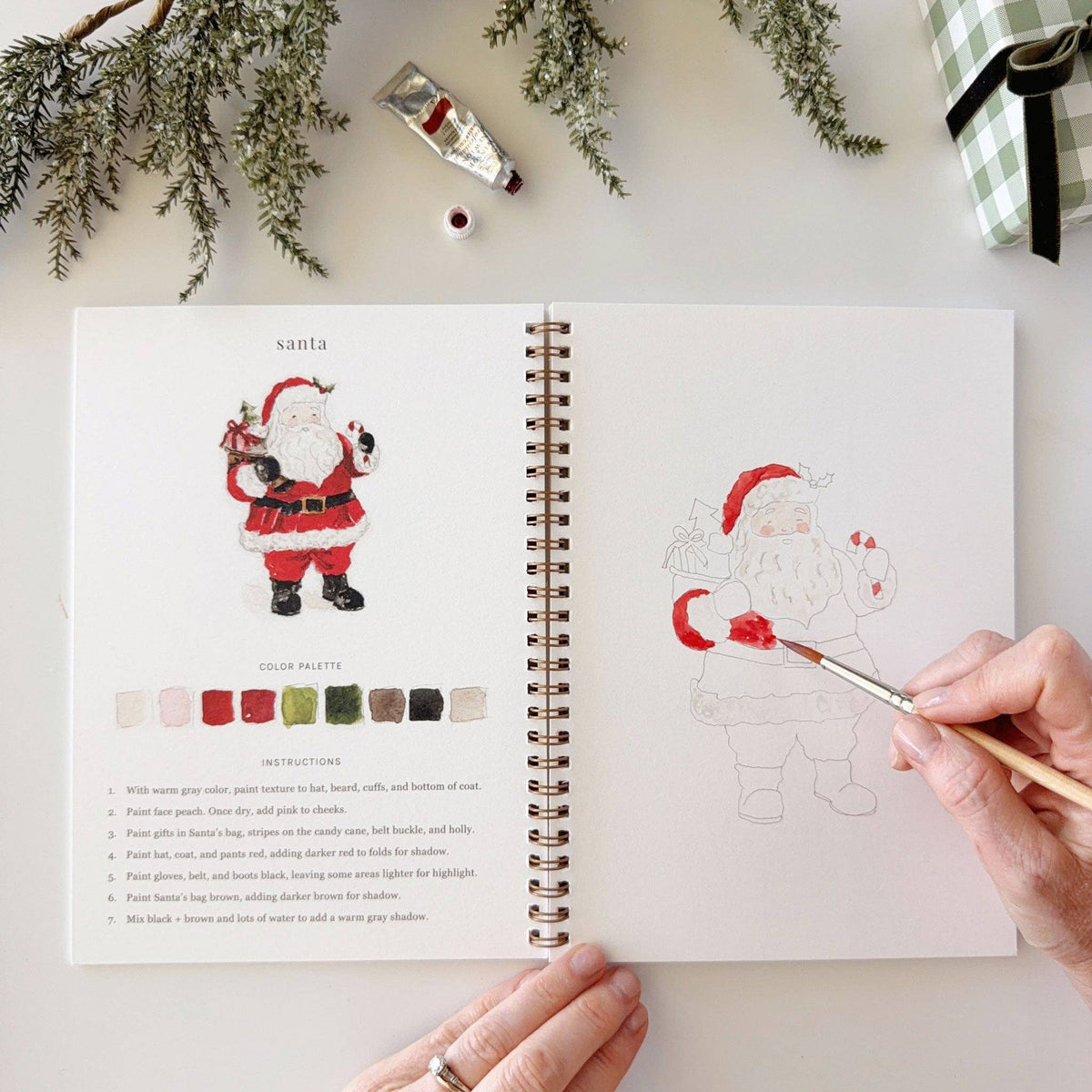 An open spiral-bound coloring book or art journal laid on a white surface, showing a Santa illustration with color palette and instructions on the left page, and a partially painted outline of Santa on the right page. 
