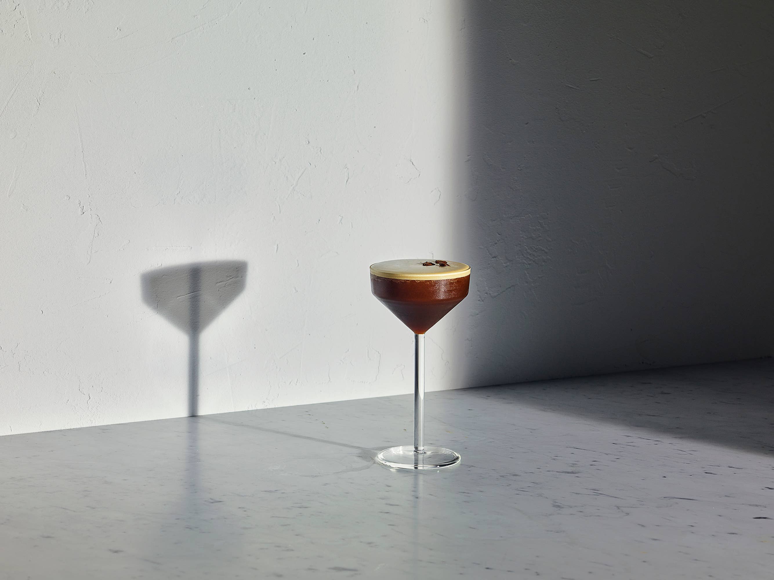 A single stemmed cocktail glass filled with layered dark brown and amber liquid, topped with a pale golden foam, standing on a smooth gray surface against a white wall with strong sunlight and shadow.
