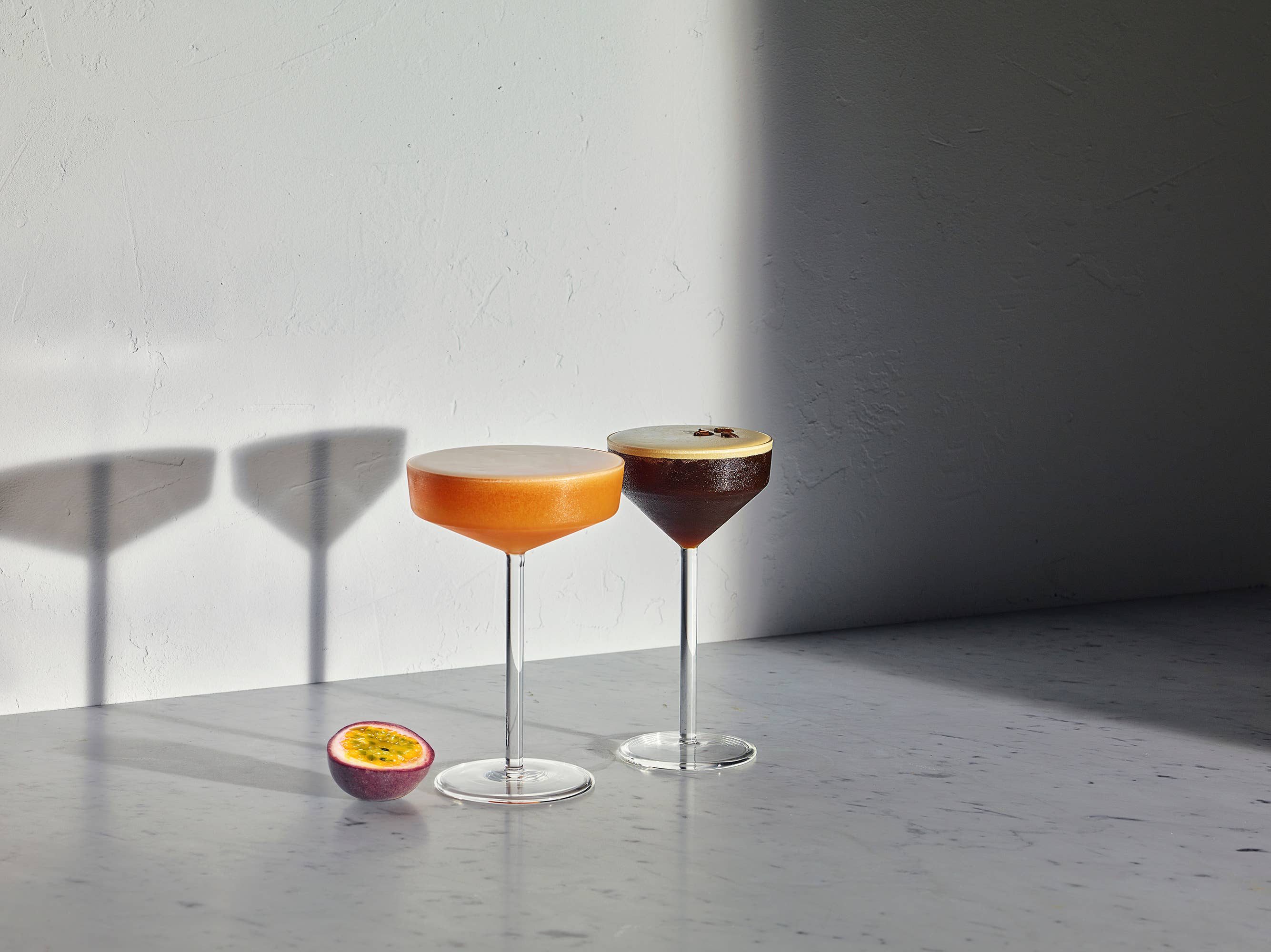 Two stemmed cocktail glasses on a gray surface against a white wall, with one filled with an orange drink and the other with a dark brown drink topped with foam. A halved passion fruit sits beside them, and strong sunlight casts shadows on the wall.