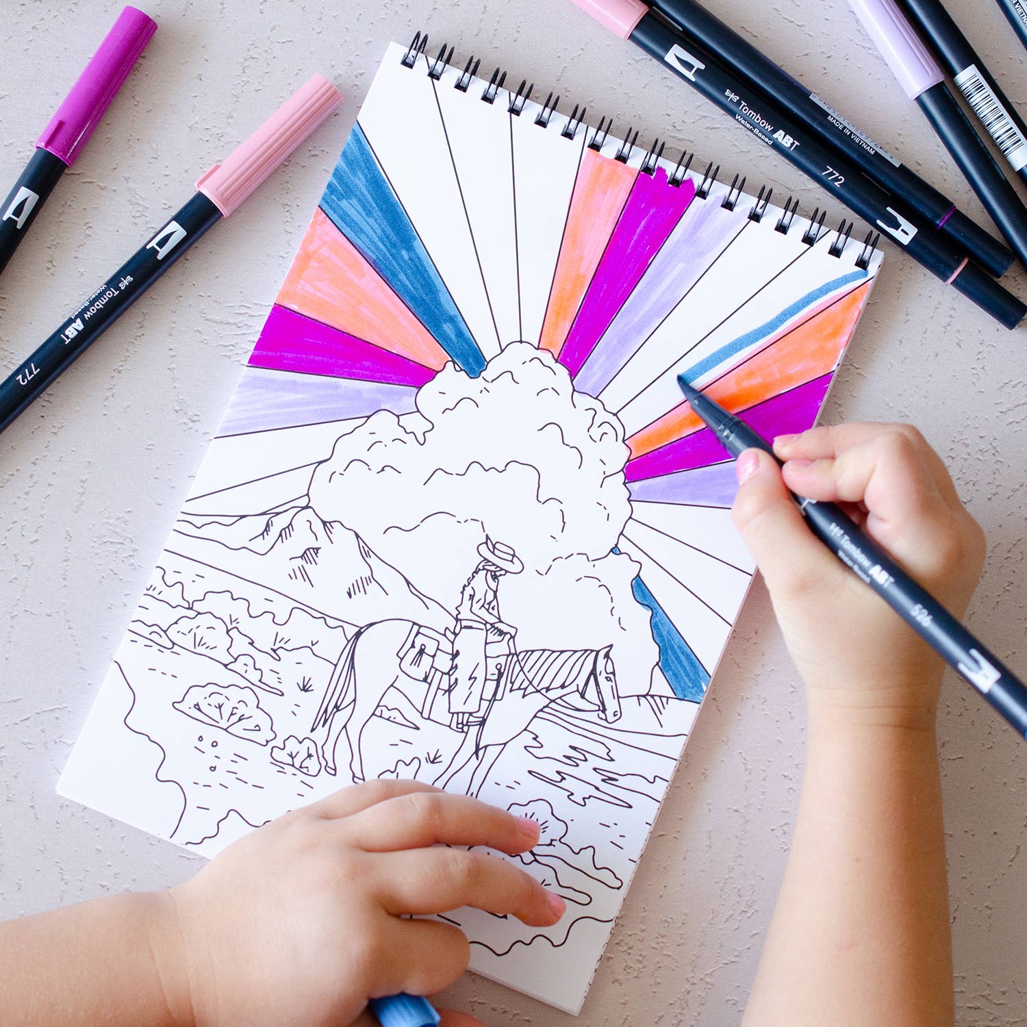Coloring book with a mountain scene and colorful markers on a light surface