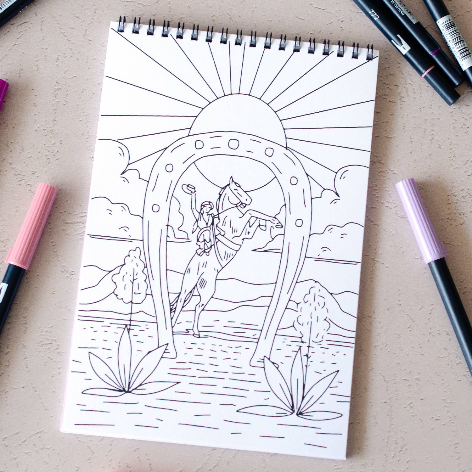Coloring book page with a design of a woman and horse, surrounded by markers on a beige surface.