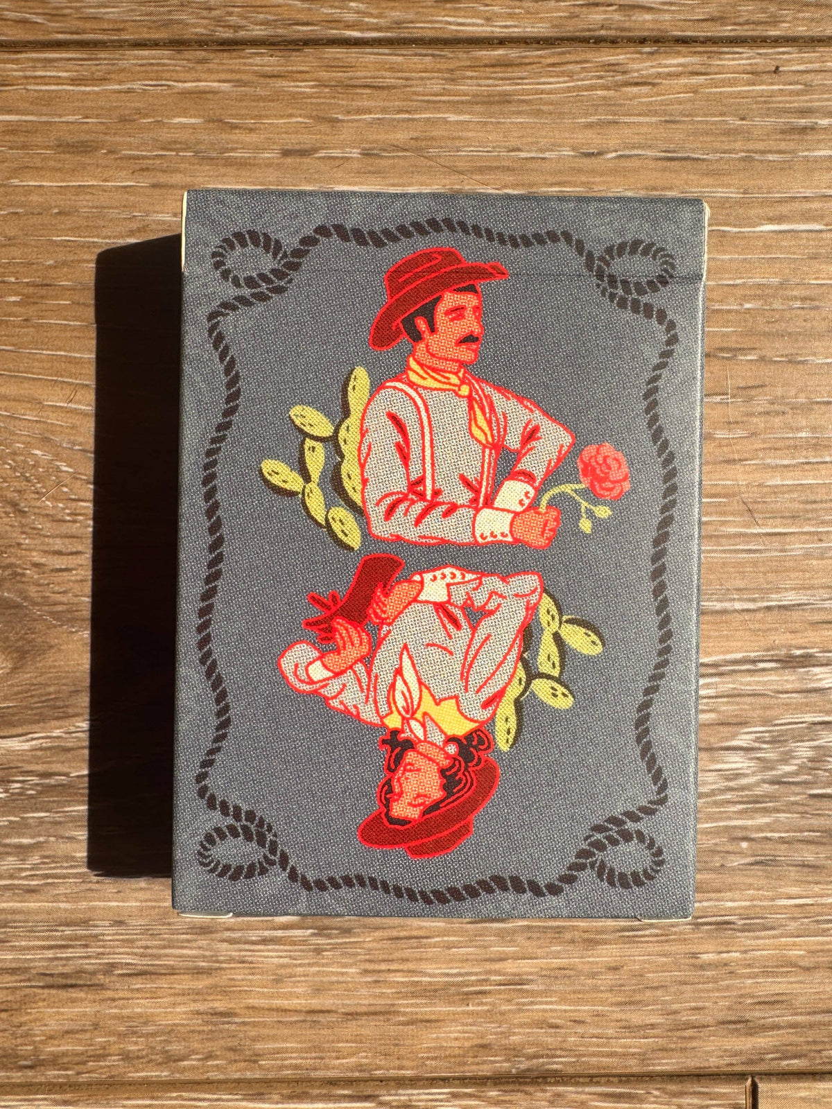 A vintage-style illustrated book or card deck box with a gray cover, featuring a red and pink cowboy illustration with yellow leaves and a black rope border.